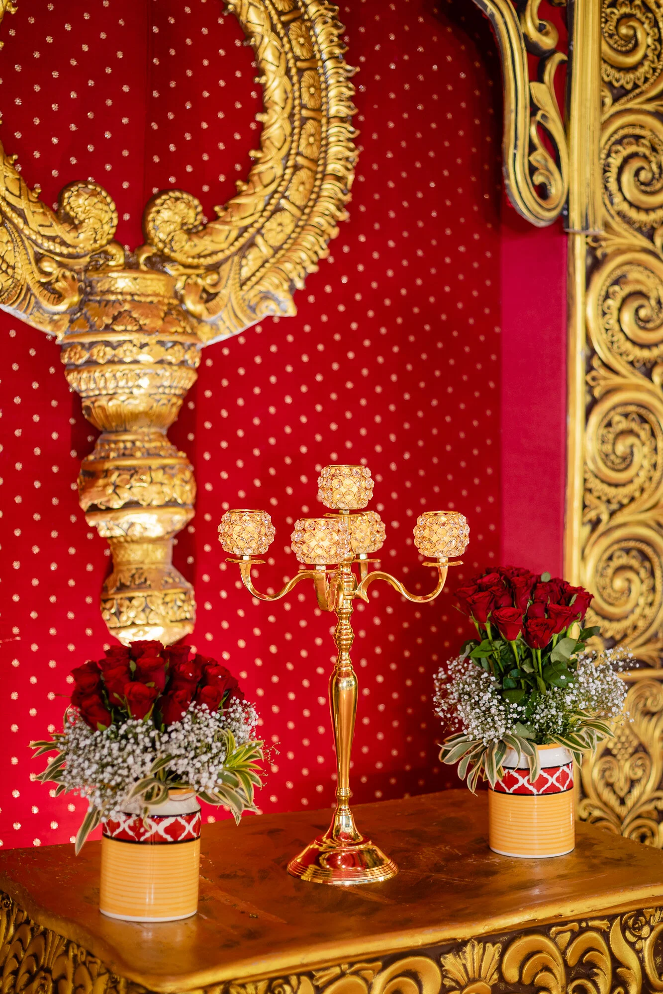 A decorative display with red roses and baby's breath in yellow and red patterned vases, a golden candelabra with crystal decorations, set against a red background with gold accents and ornate gold trim.