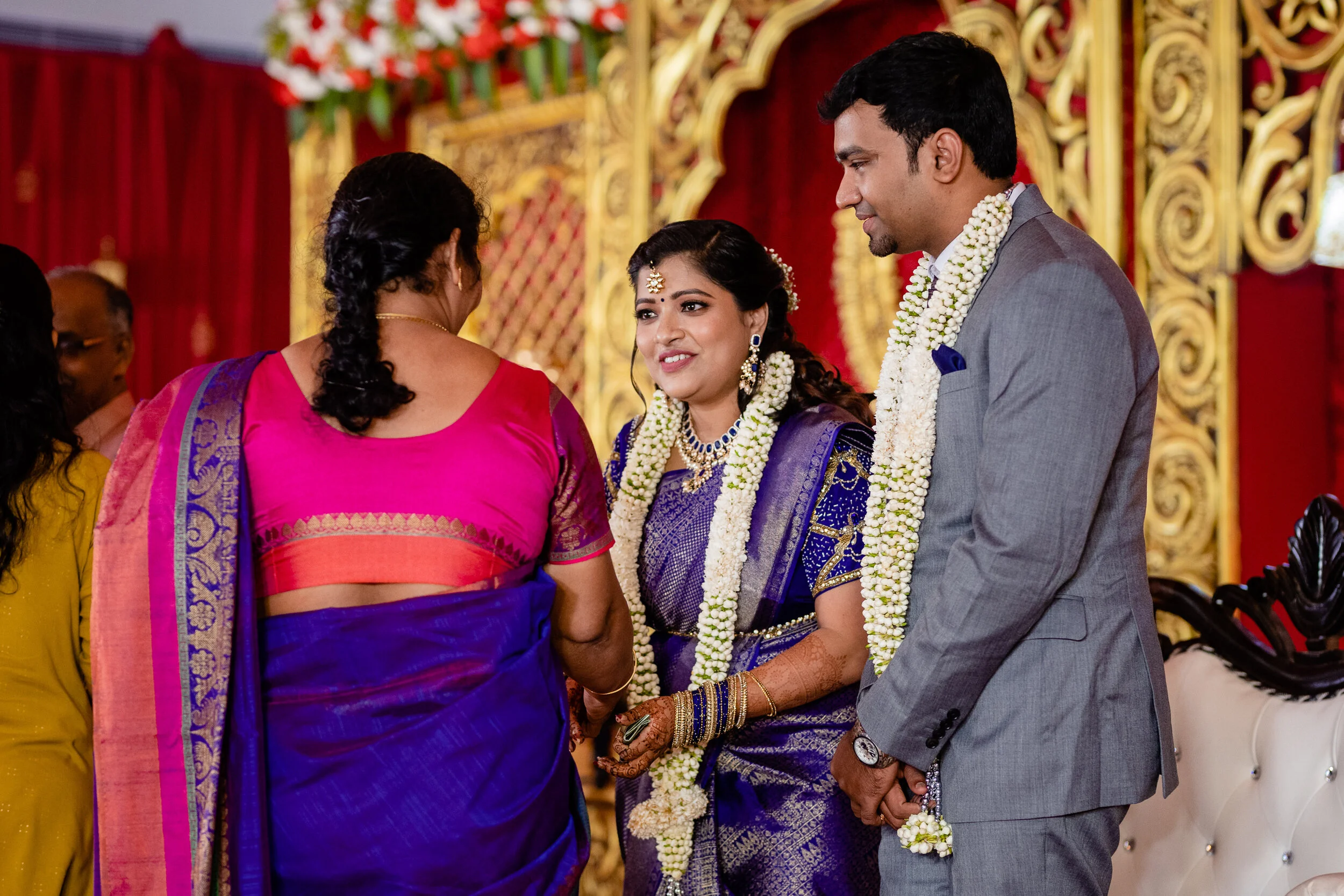 A couple dressed in traditional Indian attire participating in a wedding ceremony, standing with a woman in a pink and purple sari in a decorated venue.