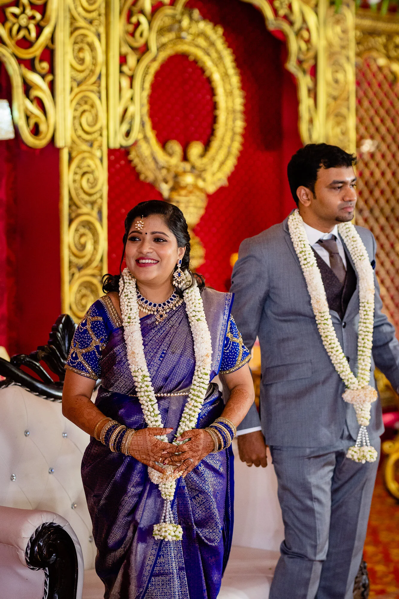 A bride and groom standing hand in hand during a wedding ceremony in India, dressed in traditional attire with floral garlands, in a decorated venue with red and gold accents.
