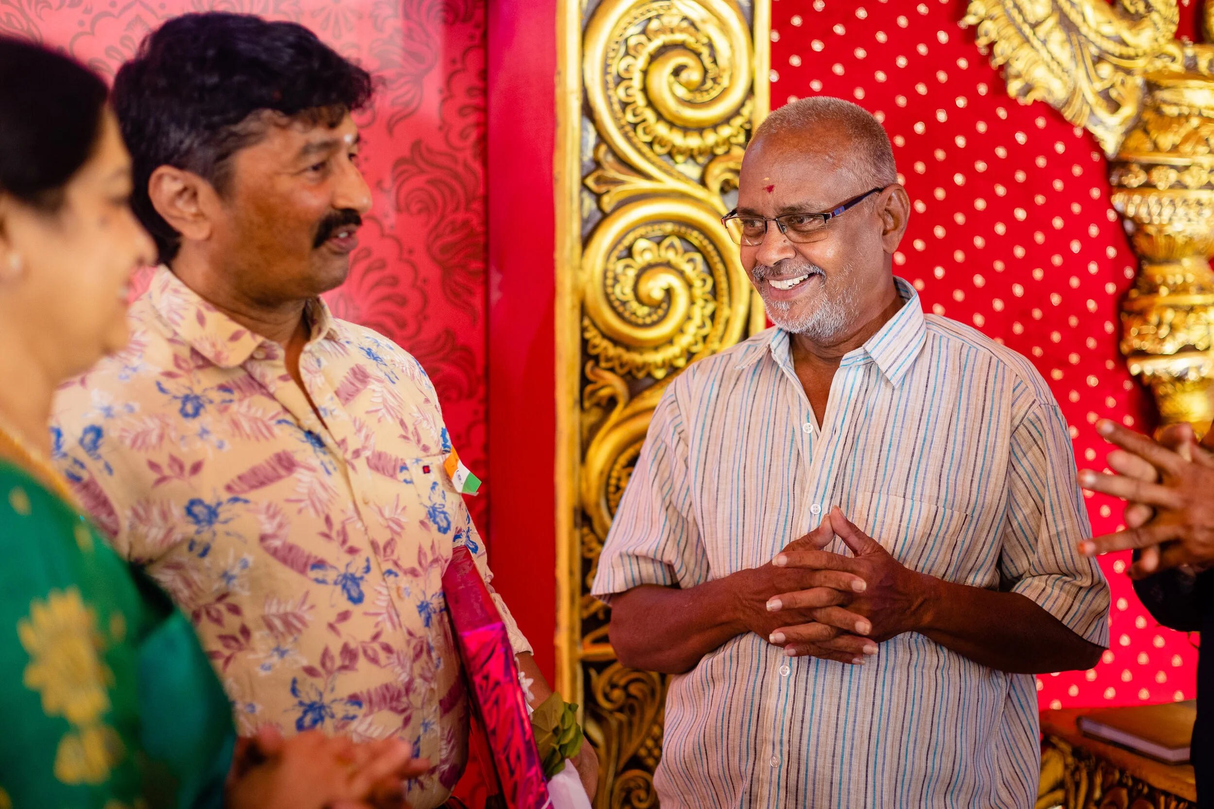 A group of people at a celebration, with the main focus on a man wearing glasses and a striped shirt, smiling and with hands clasped, standing in front of a red and gold decorated backdrop.