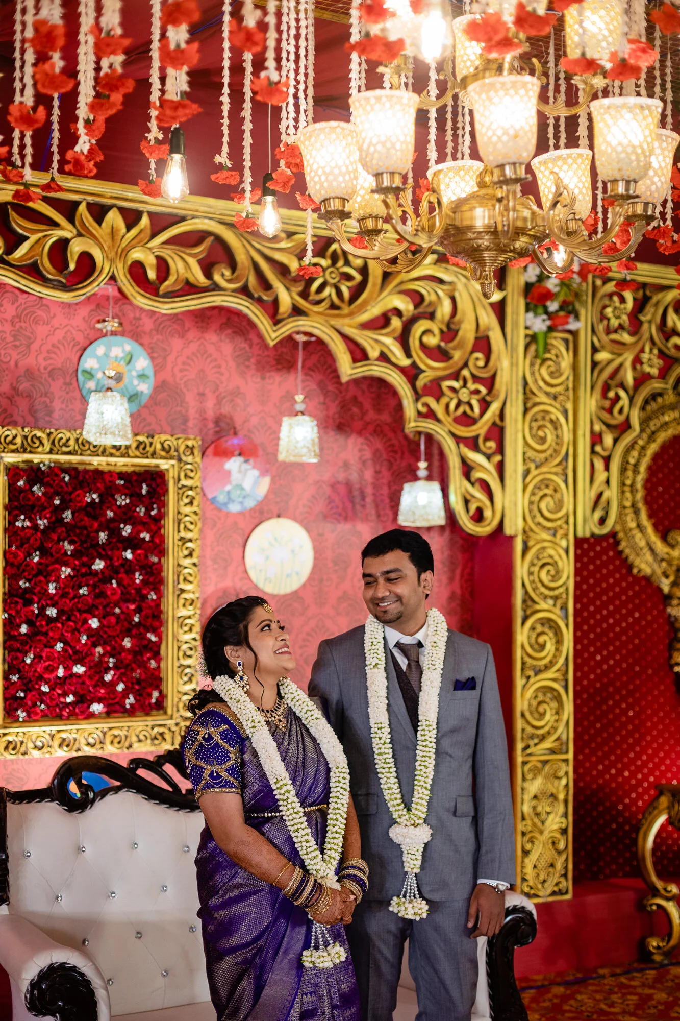 A newly married couple stands together indoors at their wedding reception or ceremony, smiling and wearing floral garlands. The woman is dressed in a purple sari with jewelry; the man is in a gray suit with a tie. The background features ornate red a