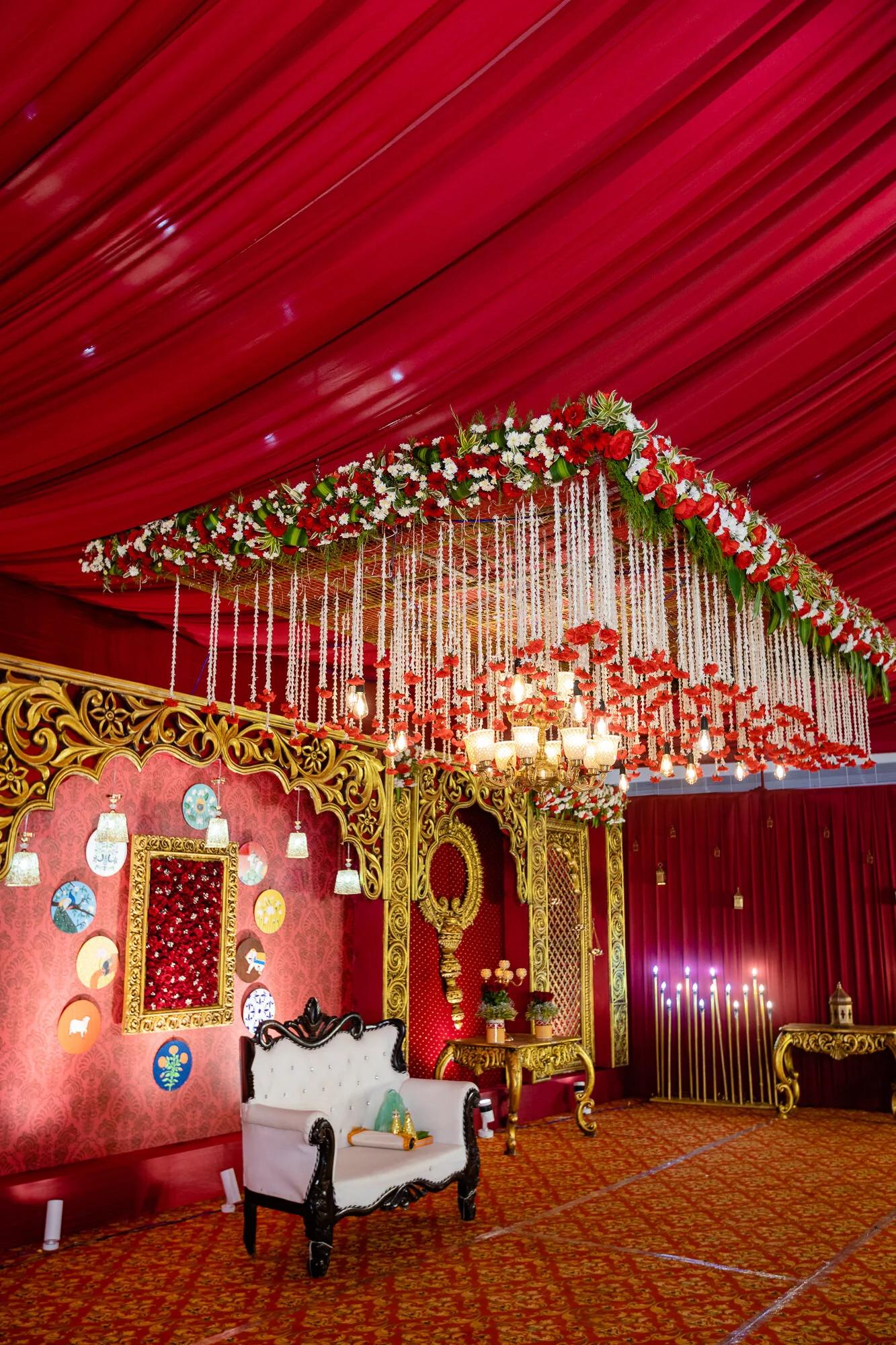 Decorated wedding hall with red and gold accents, floral chandelier, ornate furniture, red drapes, and wall art.