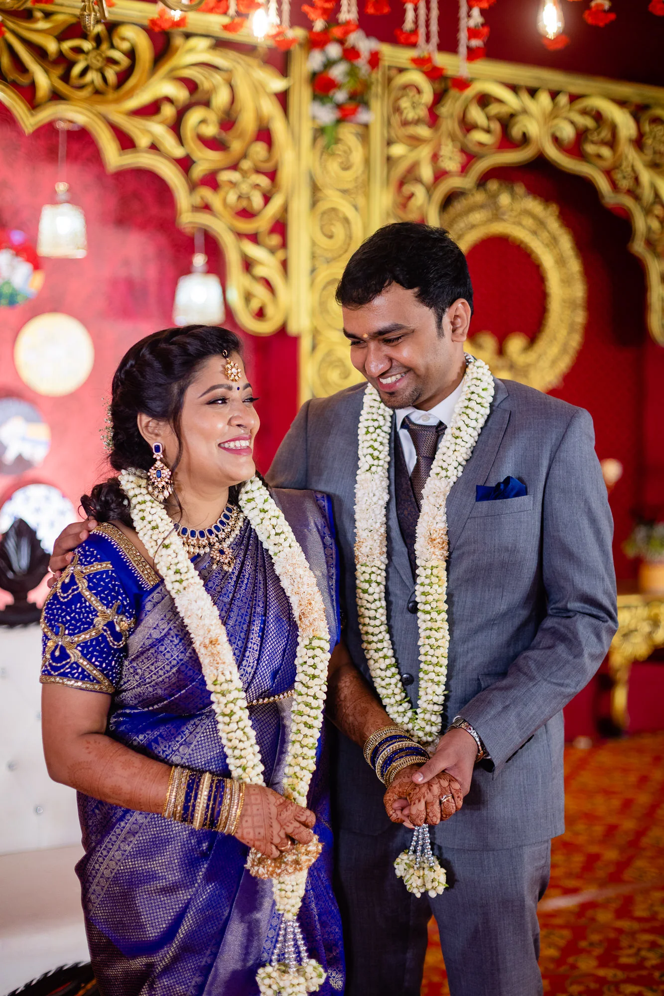 A bride and groom wearing traditional Indian wedding attire and garlands, standing together in a decorated venue, smiling and holding hands.