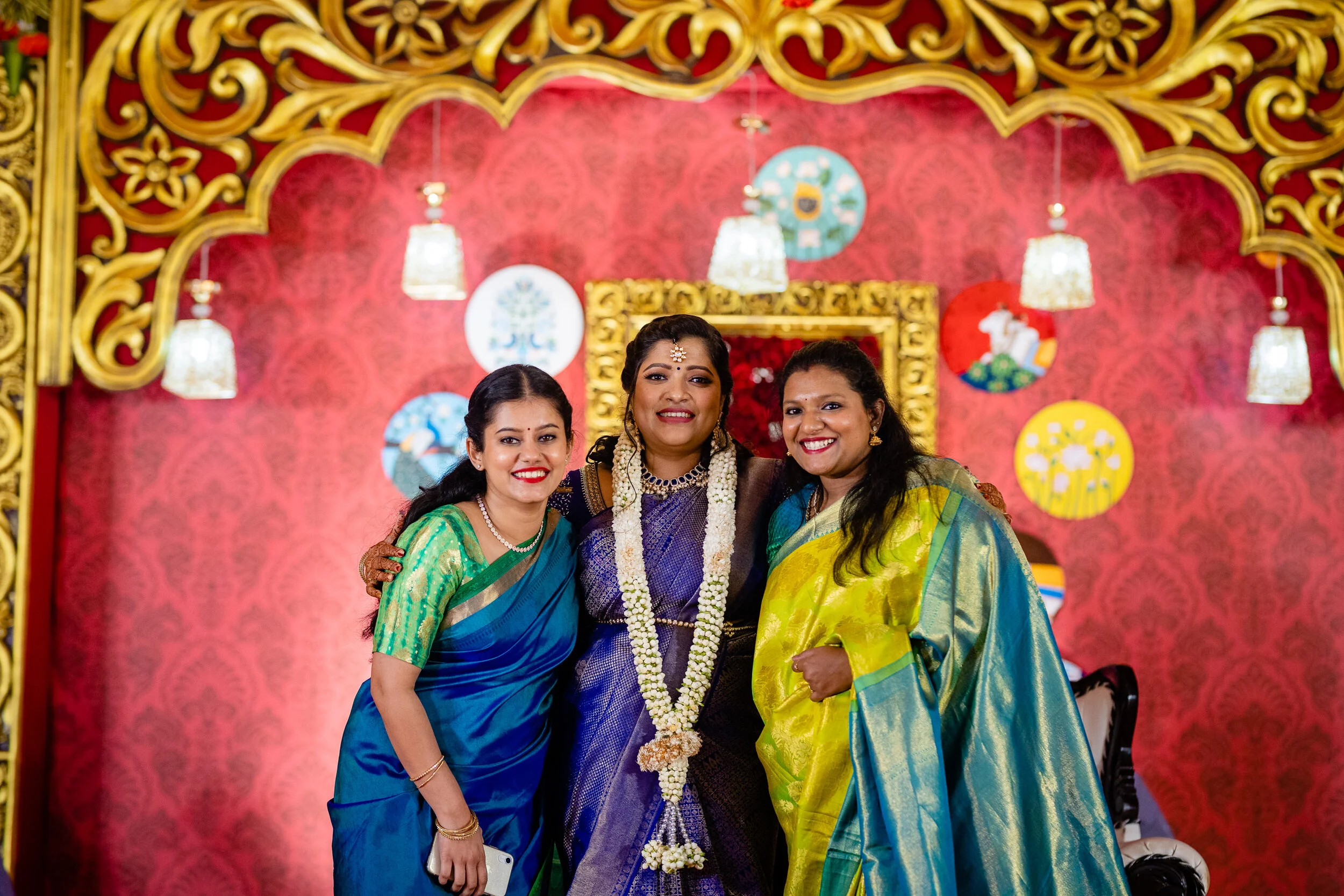 Three women dressed in colorful traditional Indian sarees smiling at a celebration, with red and gold ornate decorations in the background.