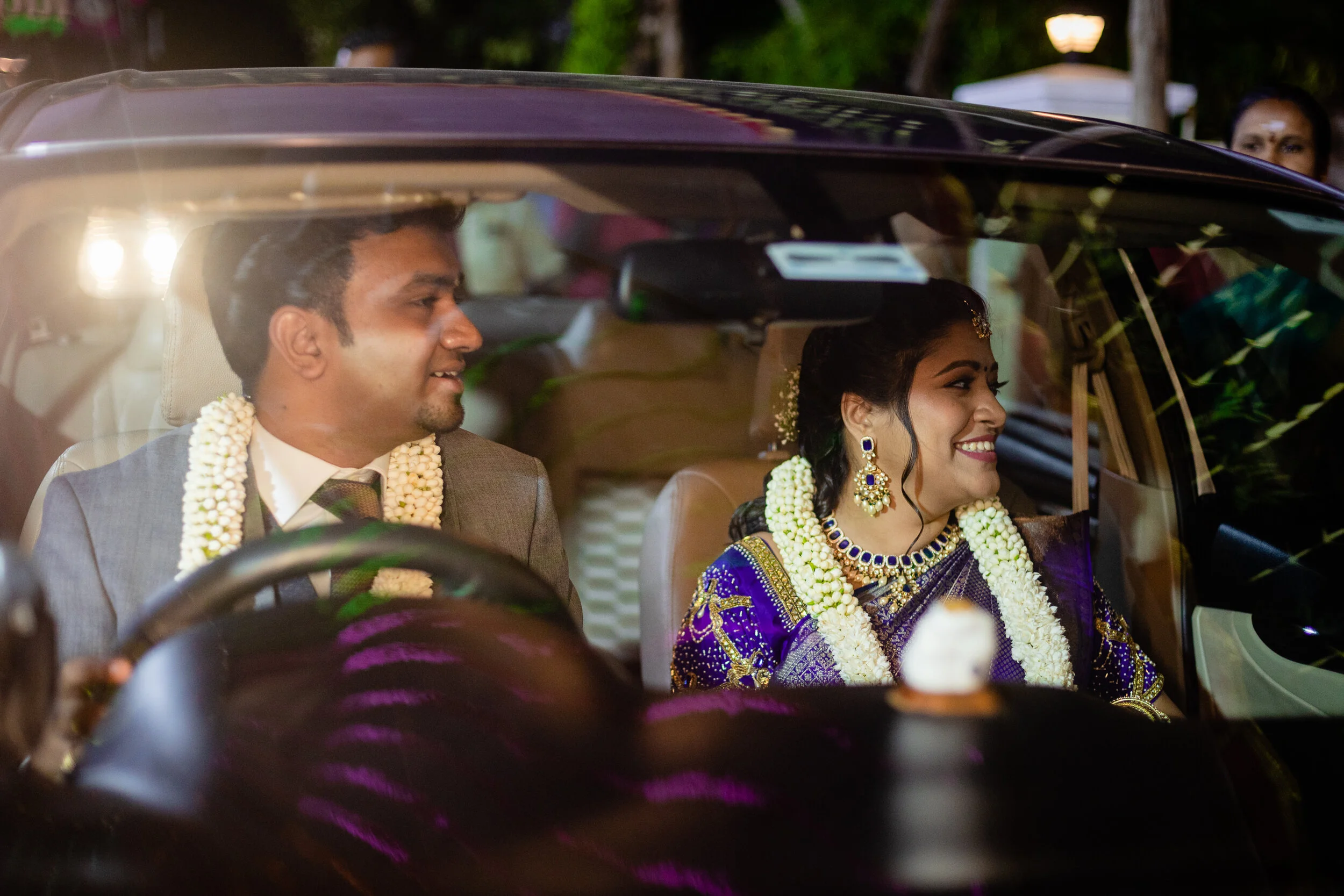 A happy bride and groom sitting in the front seat of a car decorated for a wedding, smiling at each other. The bride is wearing a purple sari with gold embroidery and jewelry, and the groom is in a suit with a flower garland around his neck.