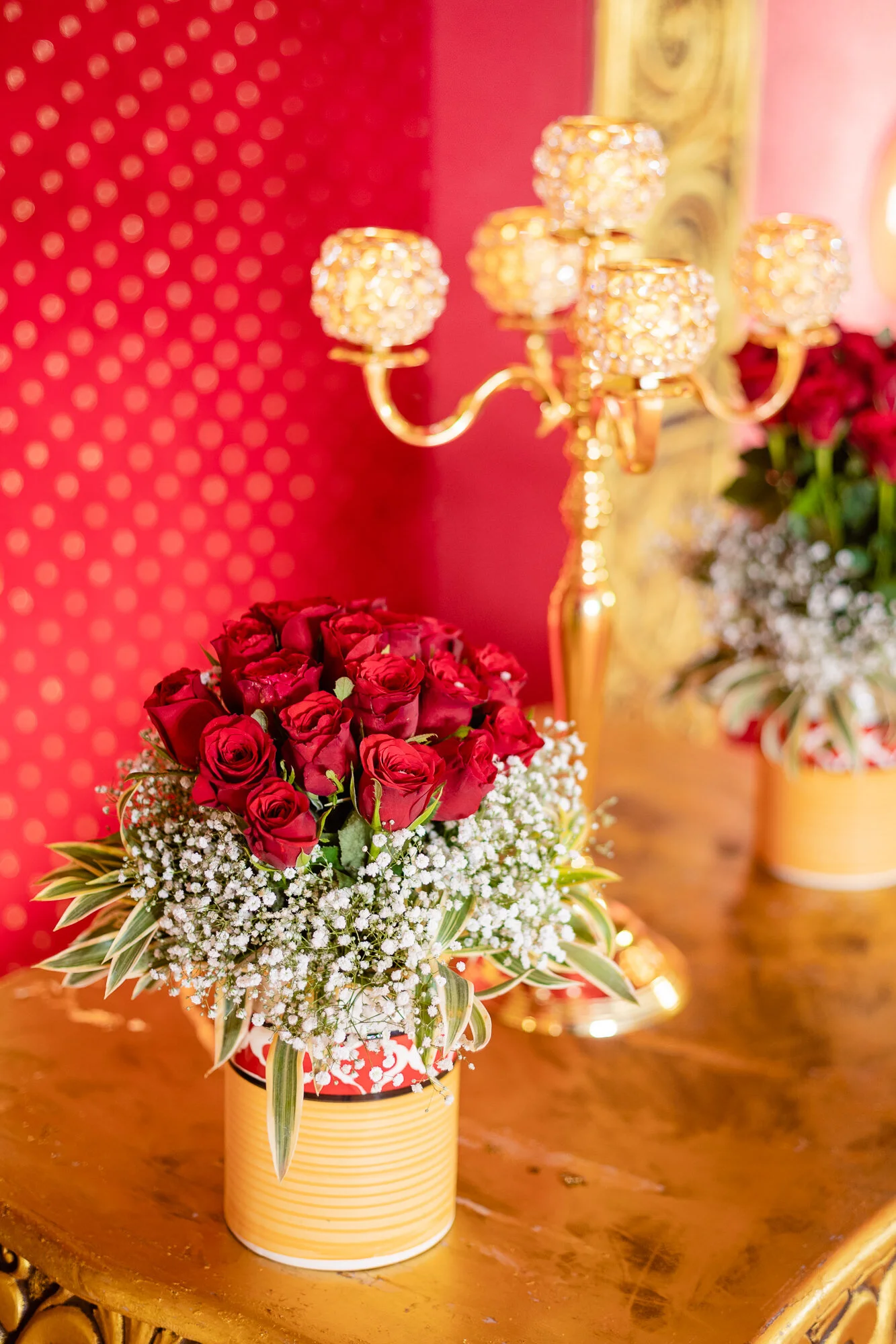 A bouquet of red roses with white baby's breath in a yellow striped vase, placed on a wooden table with a gold-colored candelabra and a red dotted background.