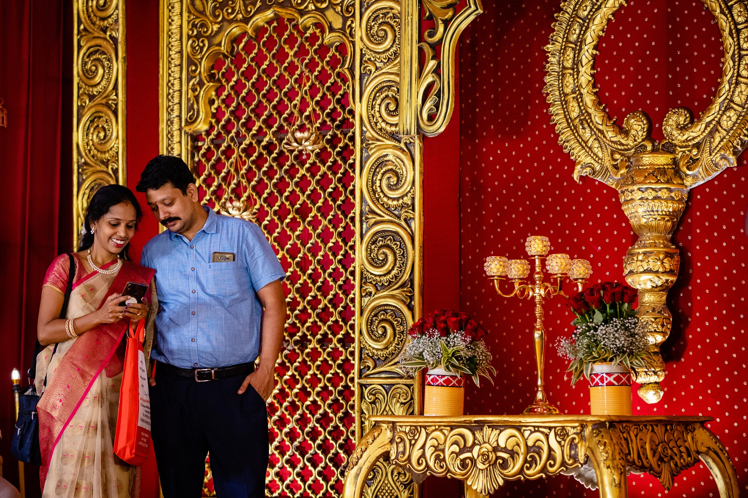 A couple dressed in traditional Indian attire looking at a phone at a decorated event with red and gold ornate decor and flower arrangements.