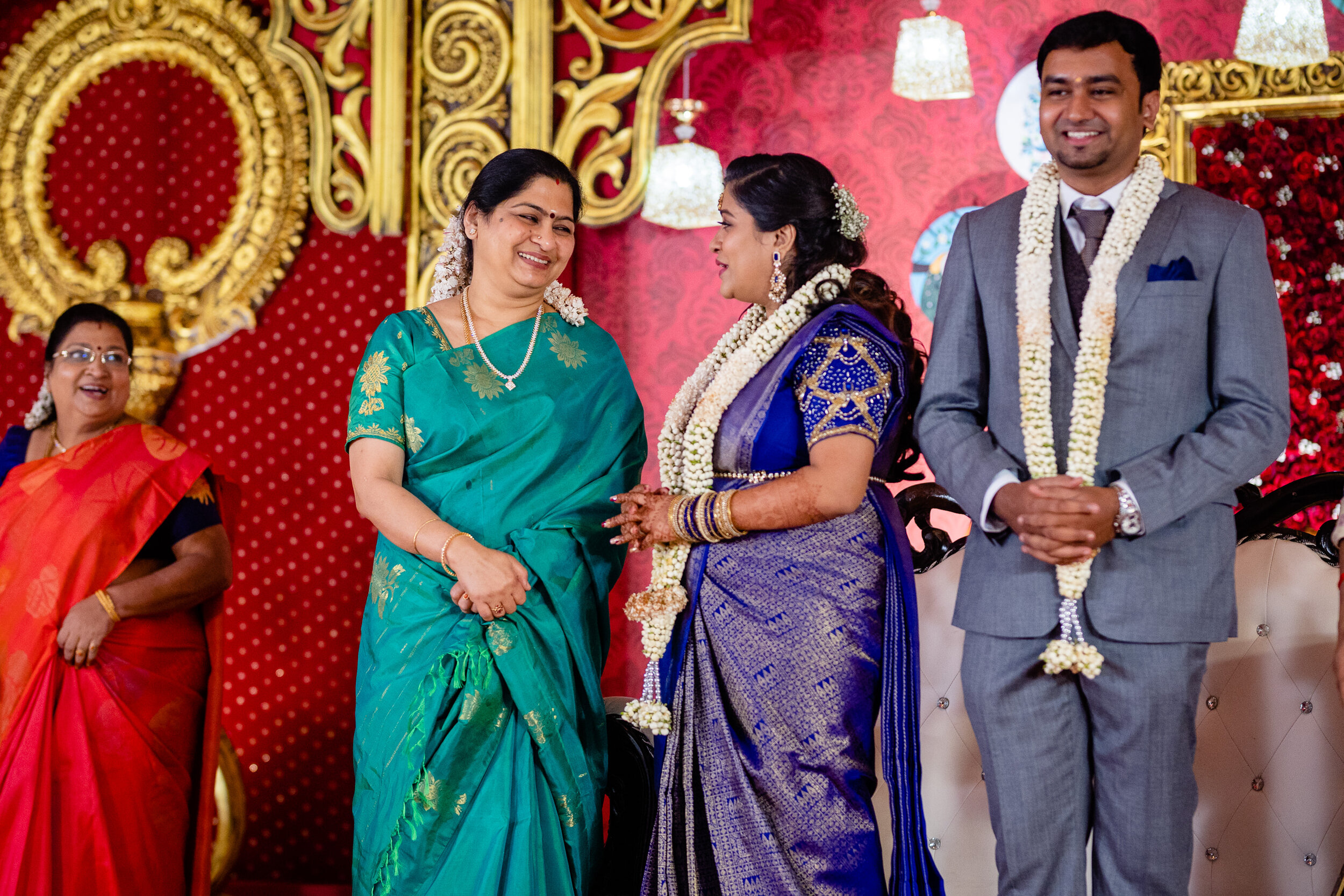 A wedding ceremony with women dressed in colorful traditional sarees and a groom in a grey suit, decorated with flower garlands, engaged in a happy moment on a decorated stage with red and gold accents.