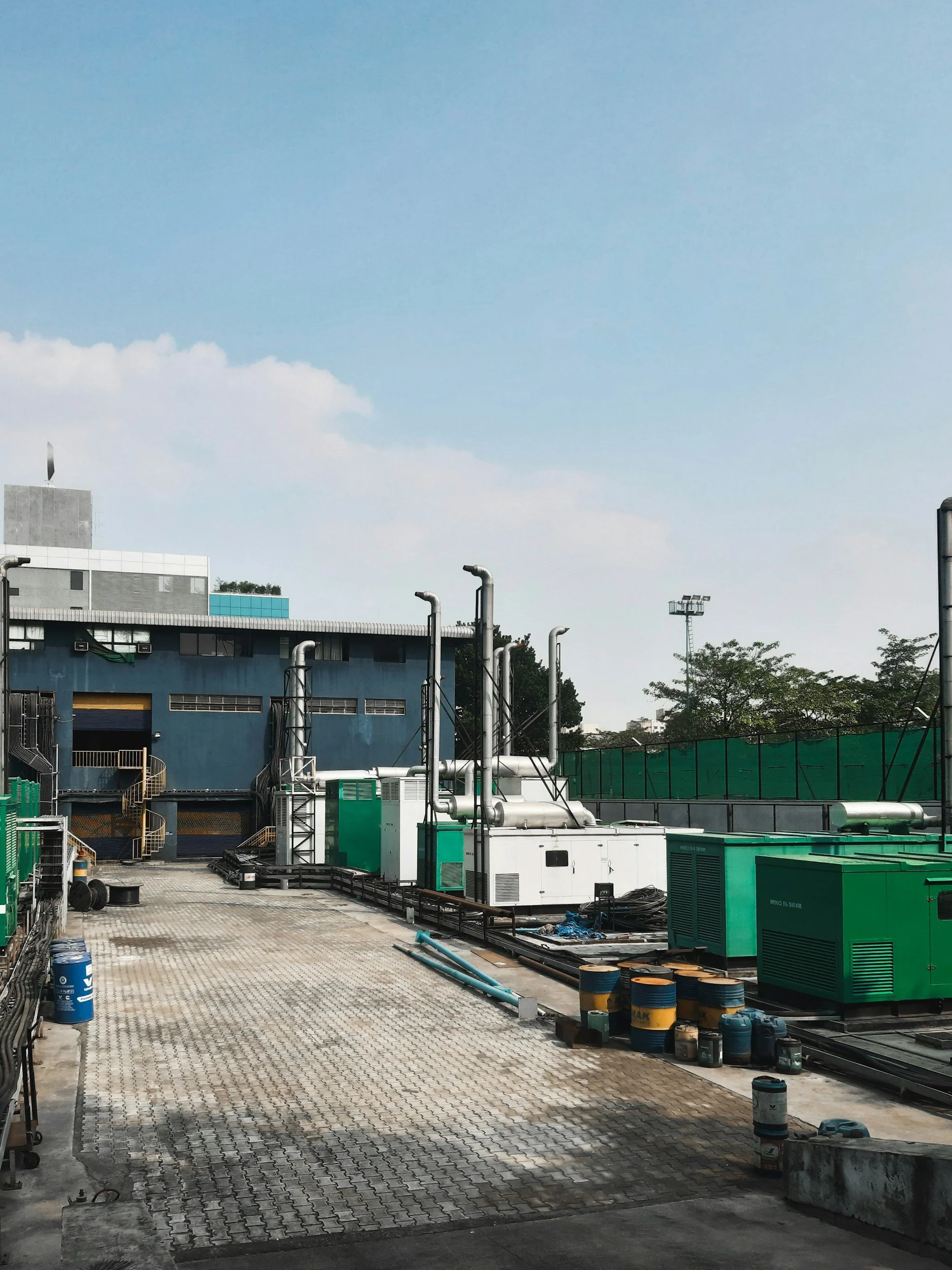 Rooftop view of an industrial area with air conditioning units, pipes, and barrels, under a blue sky with some clouds.