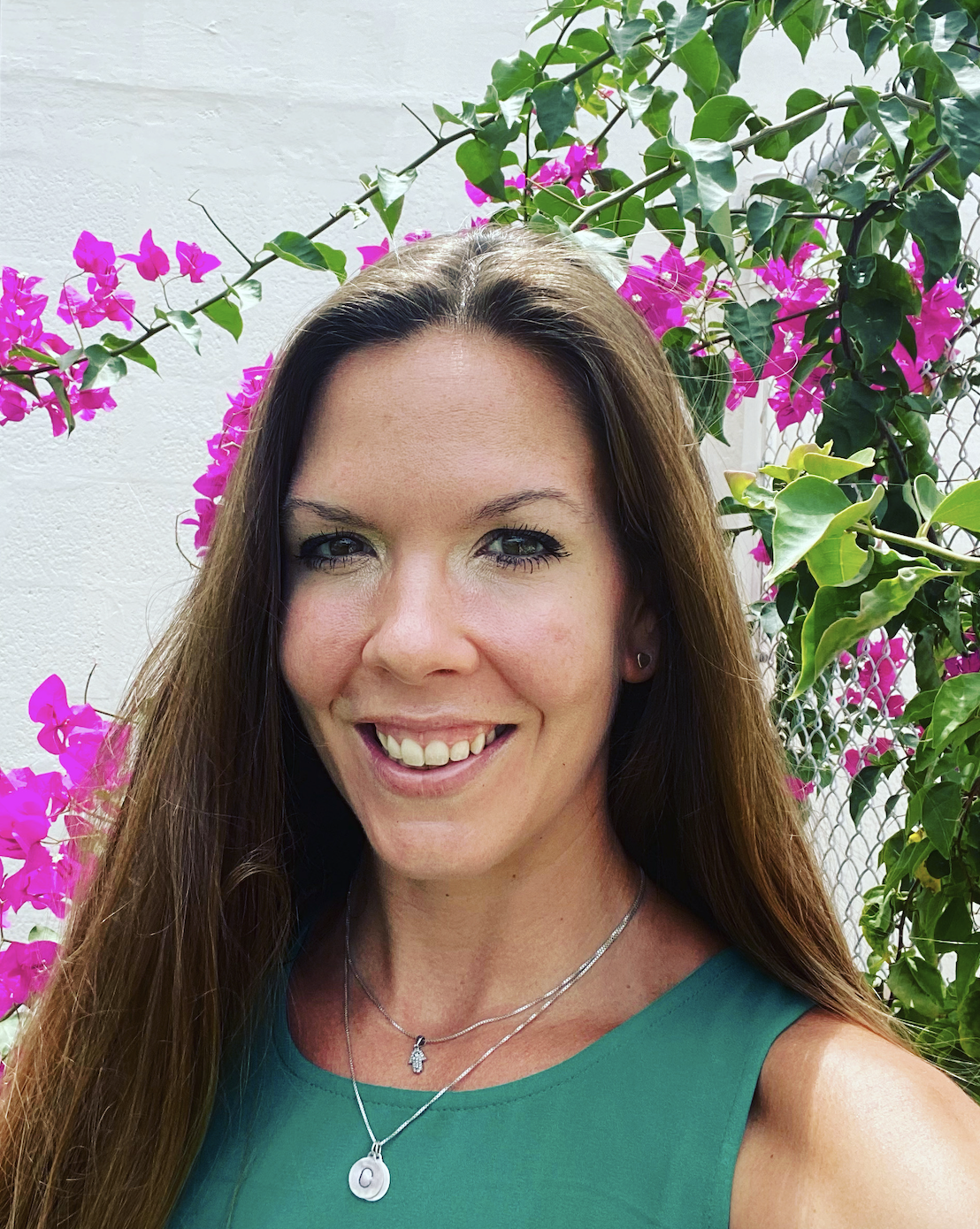 Woman with long hair smiling in front of pink flowers and green leaves.