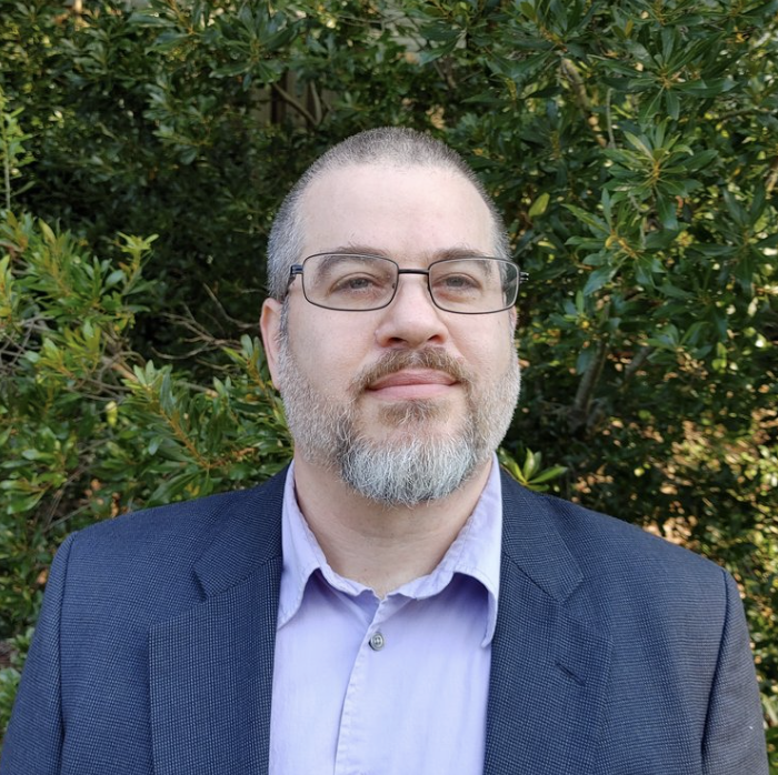Man with glasses and beard wearing a suit, standing in front of green foliage.