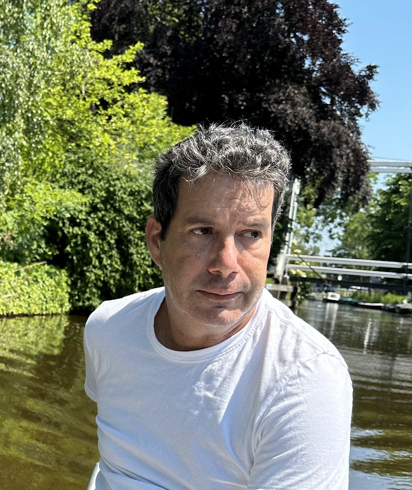Man in white shirt near a canal with lush greenery and a bridge in the background.