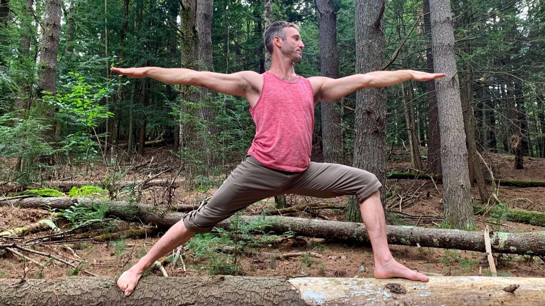 Man practicing yoga Warrior II pose on a log in a wooded area.