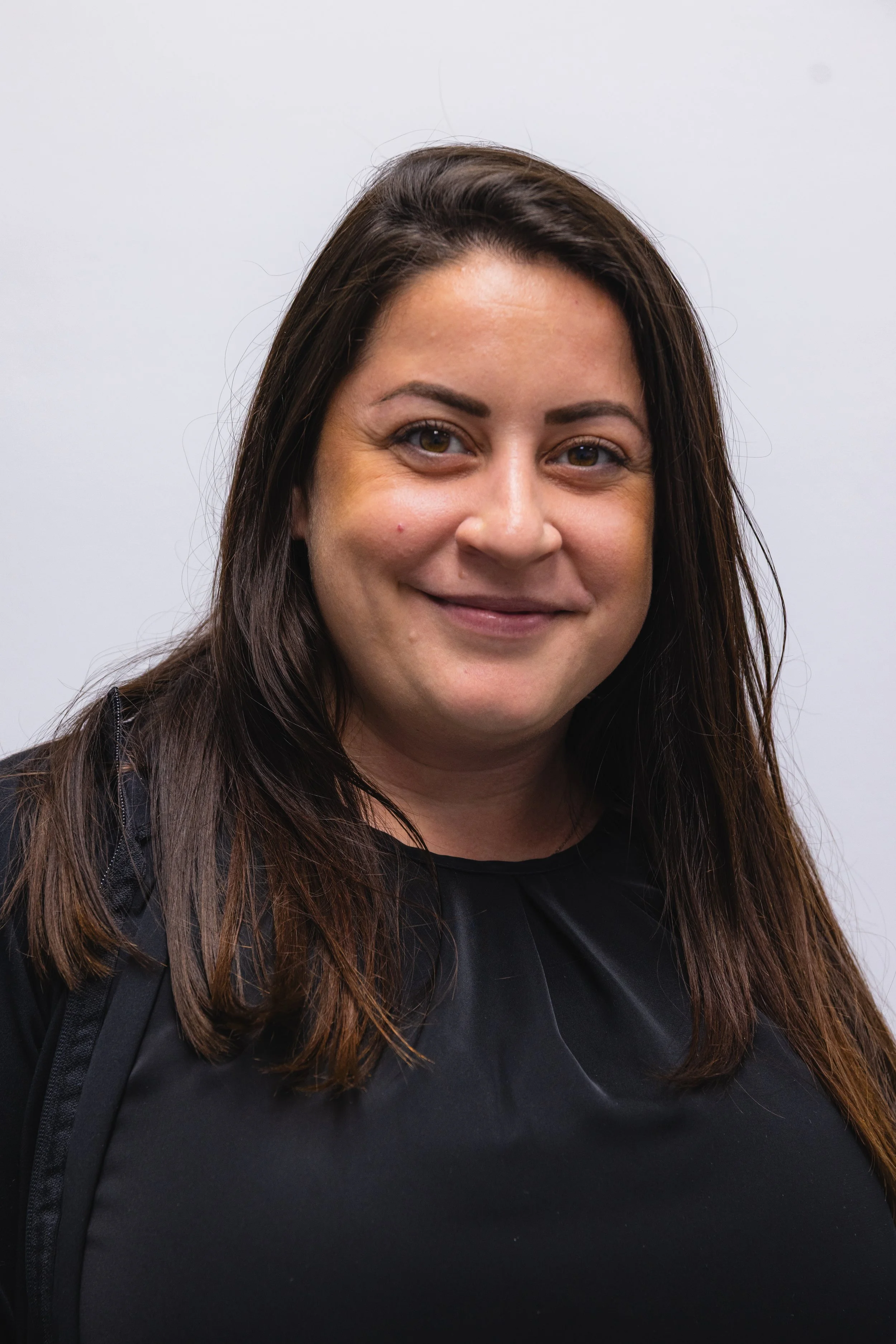 Close-up of a smiling woman with long dark hair, wearing a black top and backpack against a plain white background.