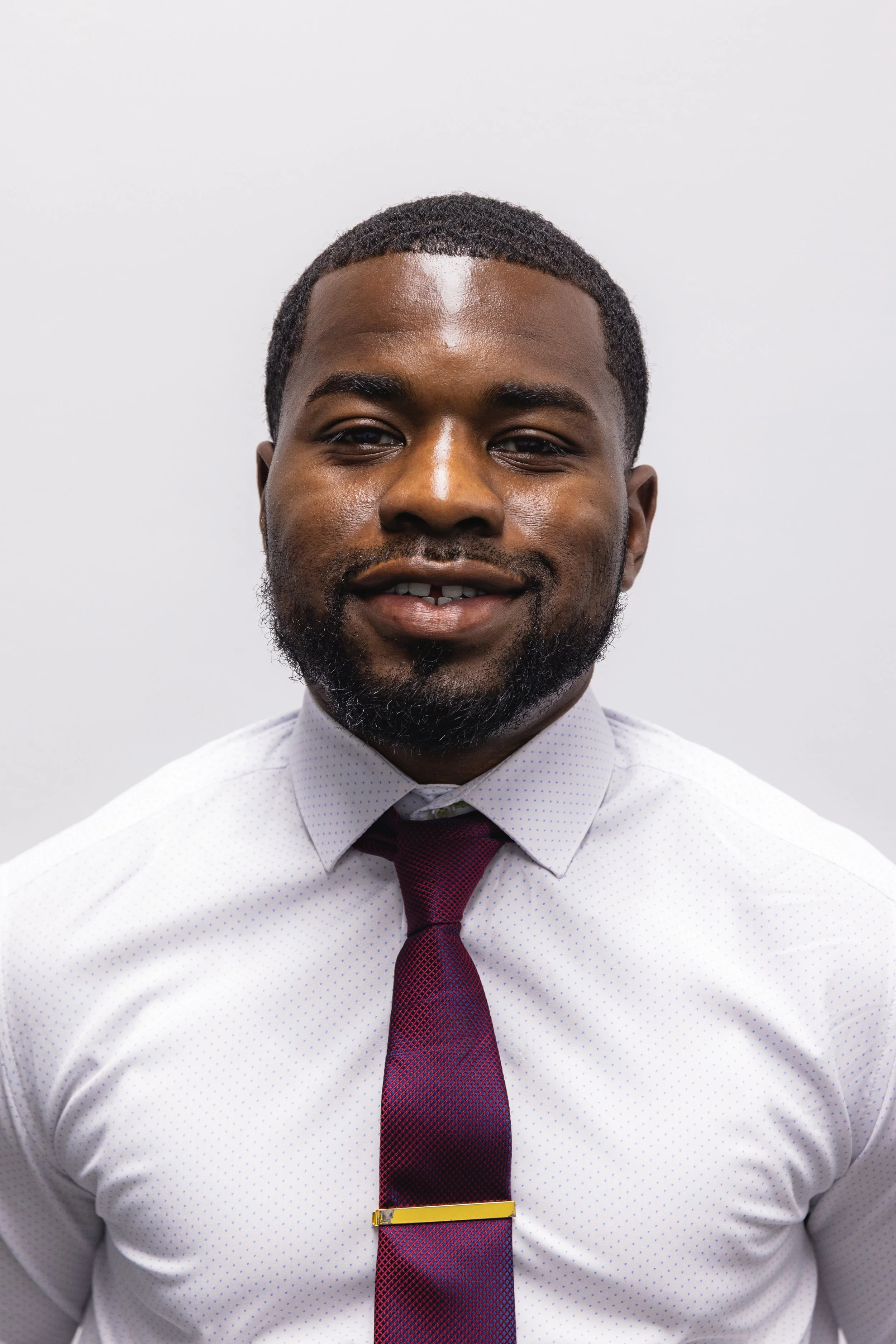 A young man with short curly hair and a beard smiling, wearing a white dress shirt with tiny blue dots and a red and blue tie, standing against a plain light gray background.