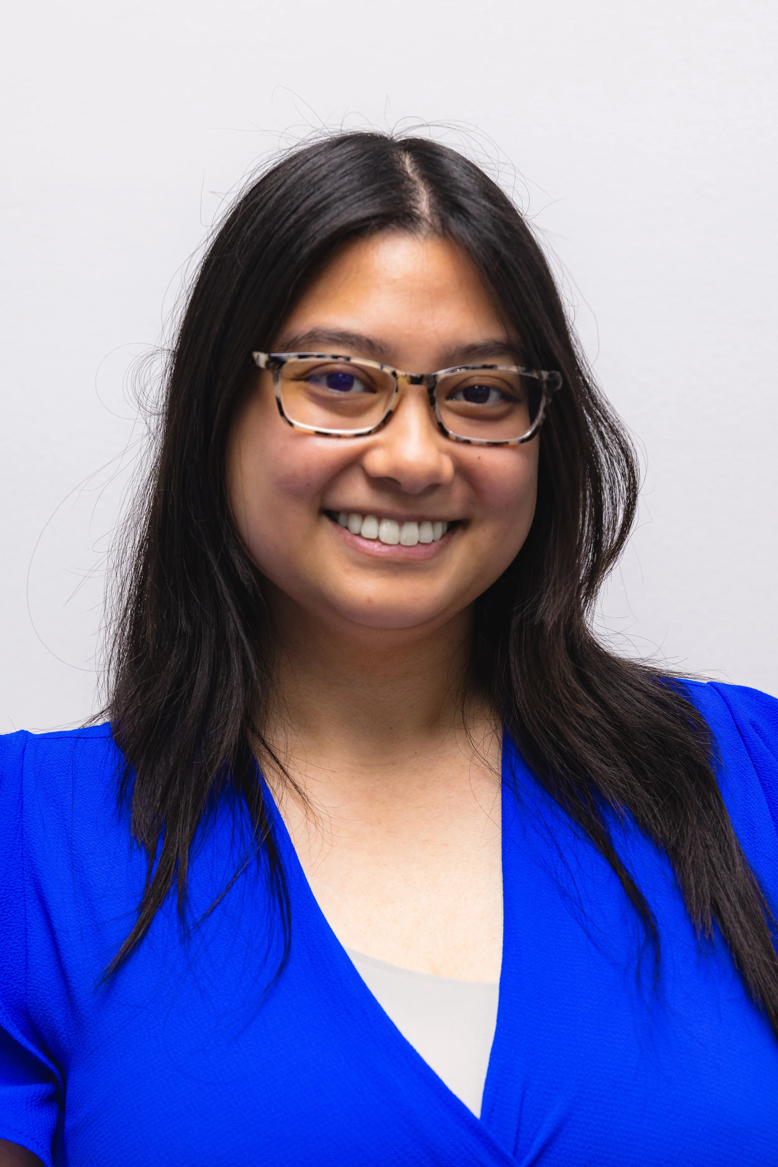 A woman with long black hair and glasses, smiling, wearing a royal blue top over a white shirt, standing in front of a plain light gray background.