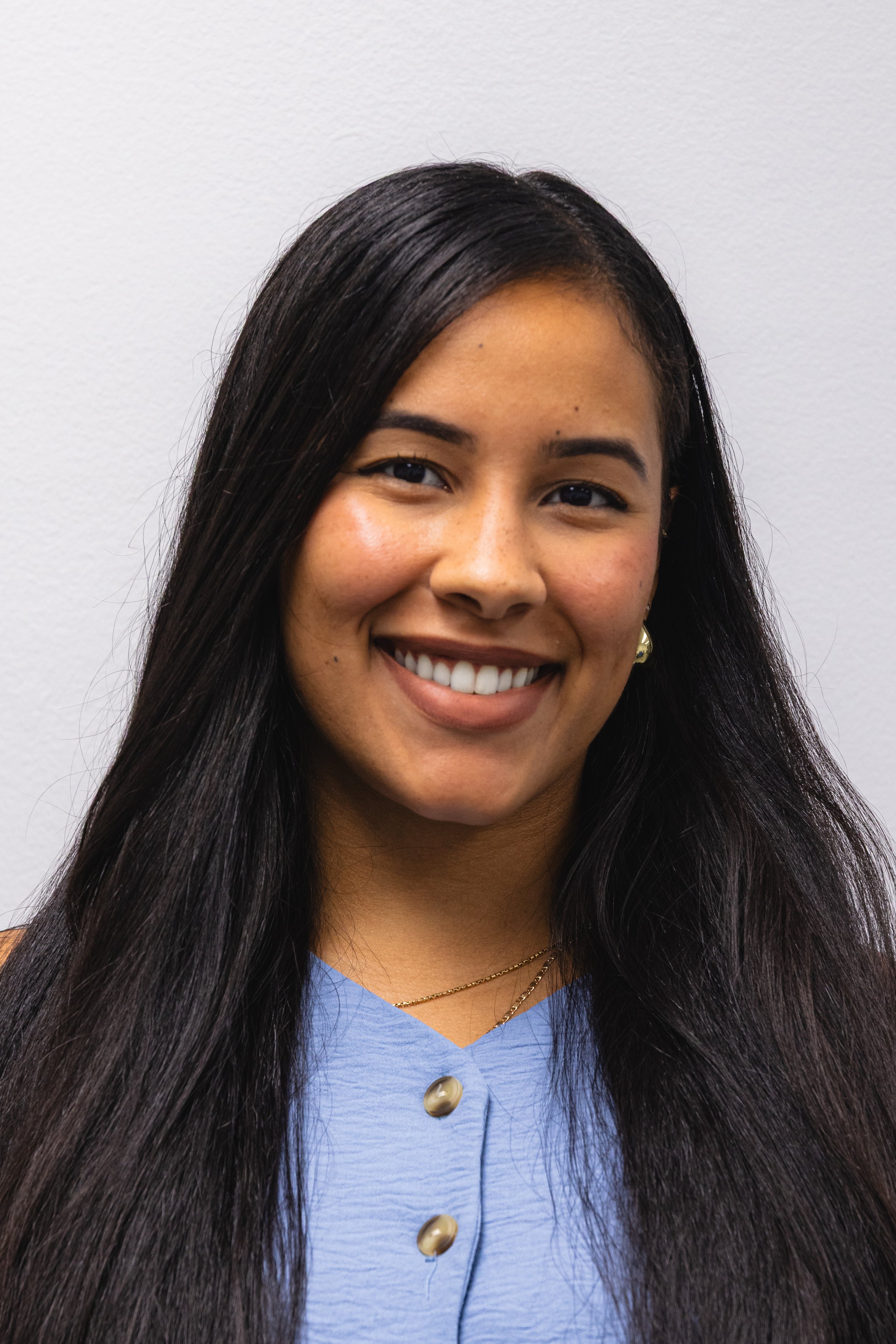 Close-up of a woman with long dark hair, smiling, wearing a blue shirt with gold buttons, a necklace, and earrings, against a plain white wall.
