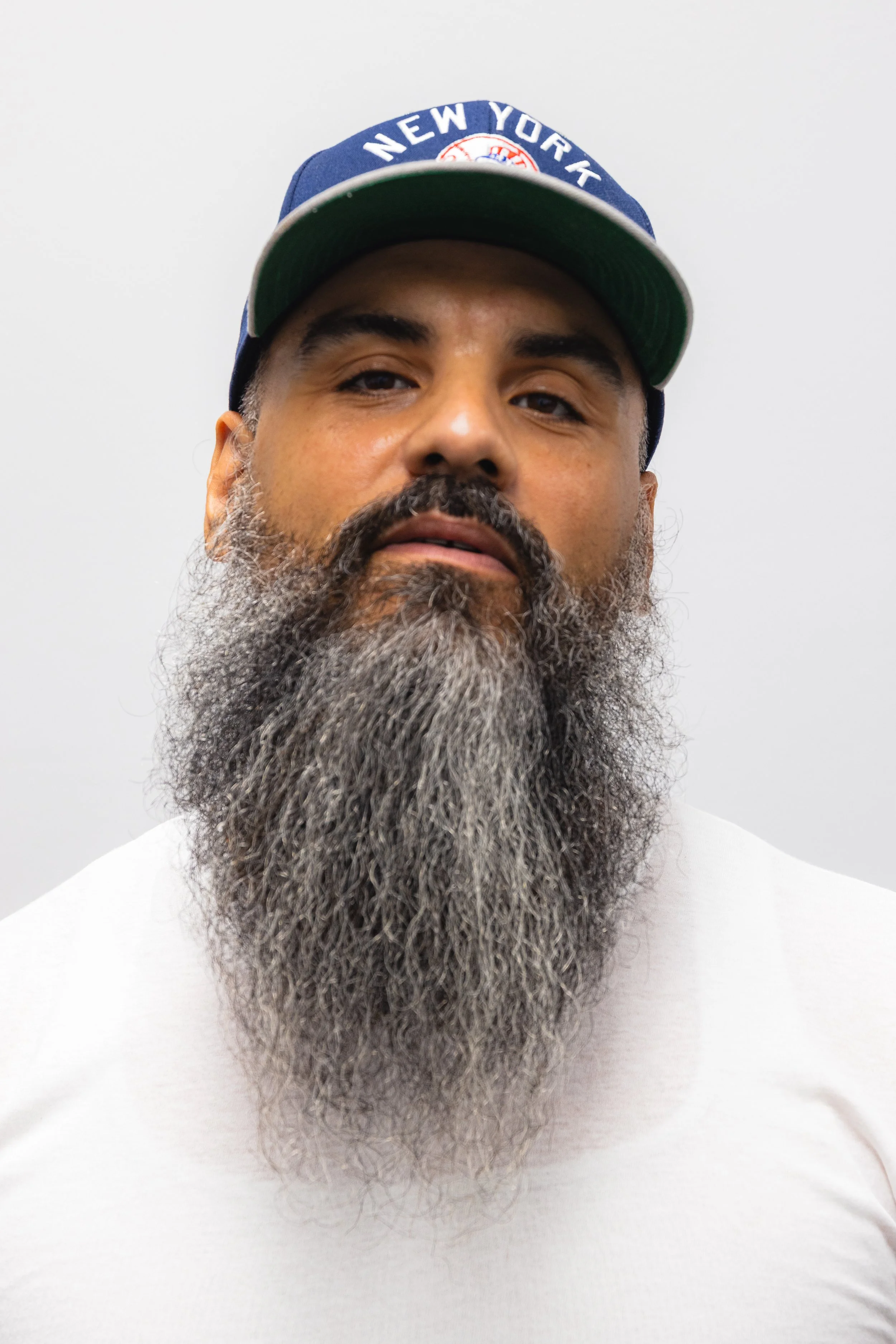 Close-up of a man with a gray beard and mustache wearing a blue cap with 'NEW YORK' written on it, against a plain white background.