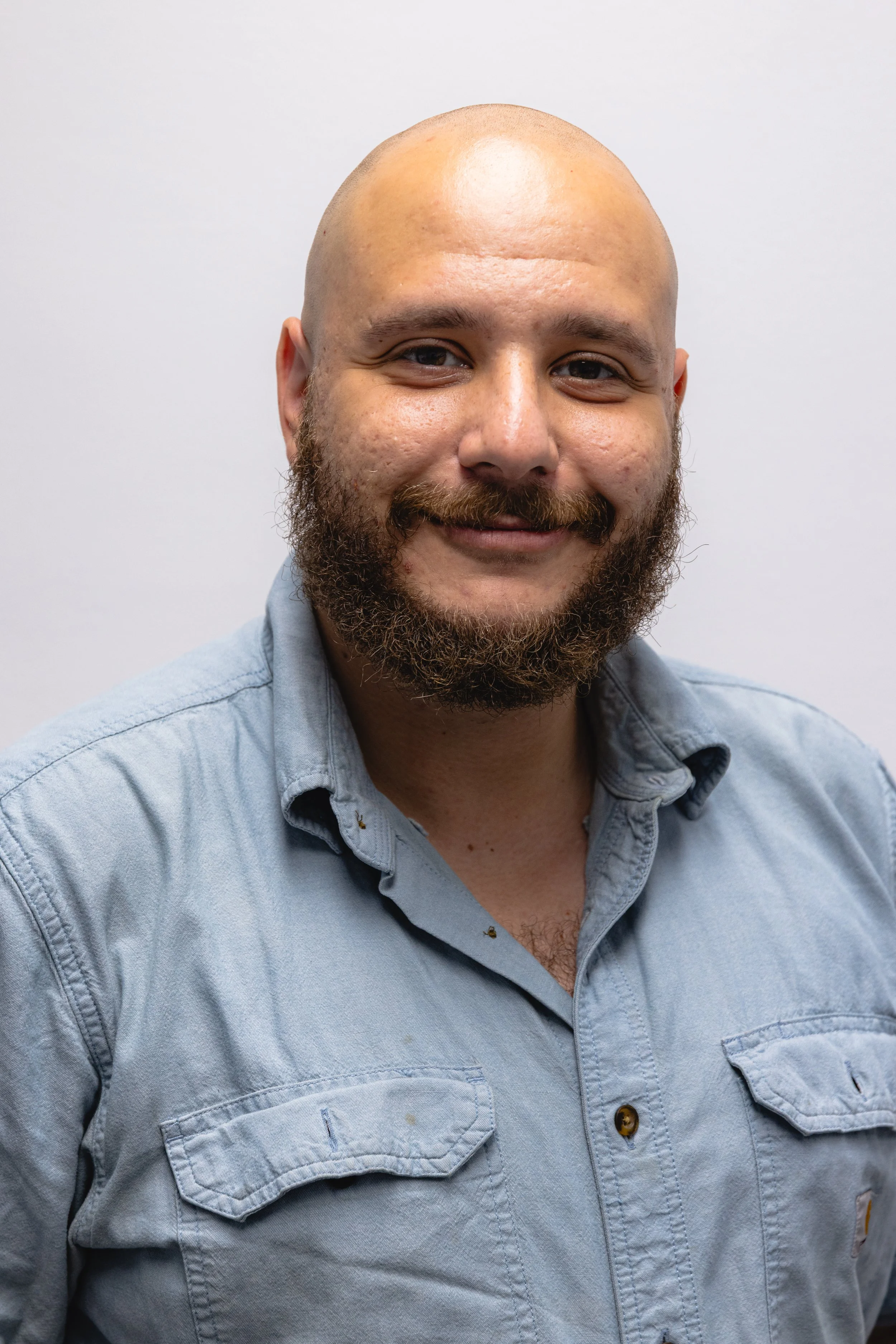 A smiling man with a beard, wearing a light blue button-up shirt, standing against a plain white wall.