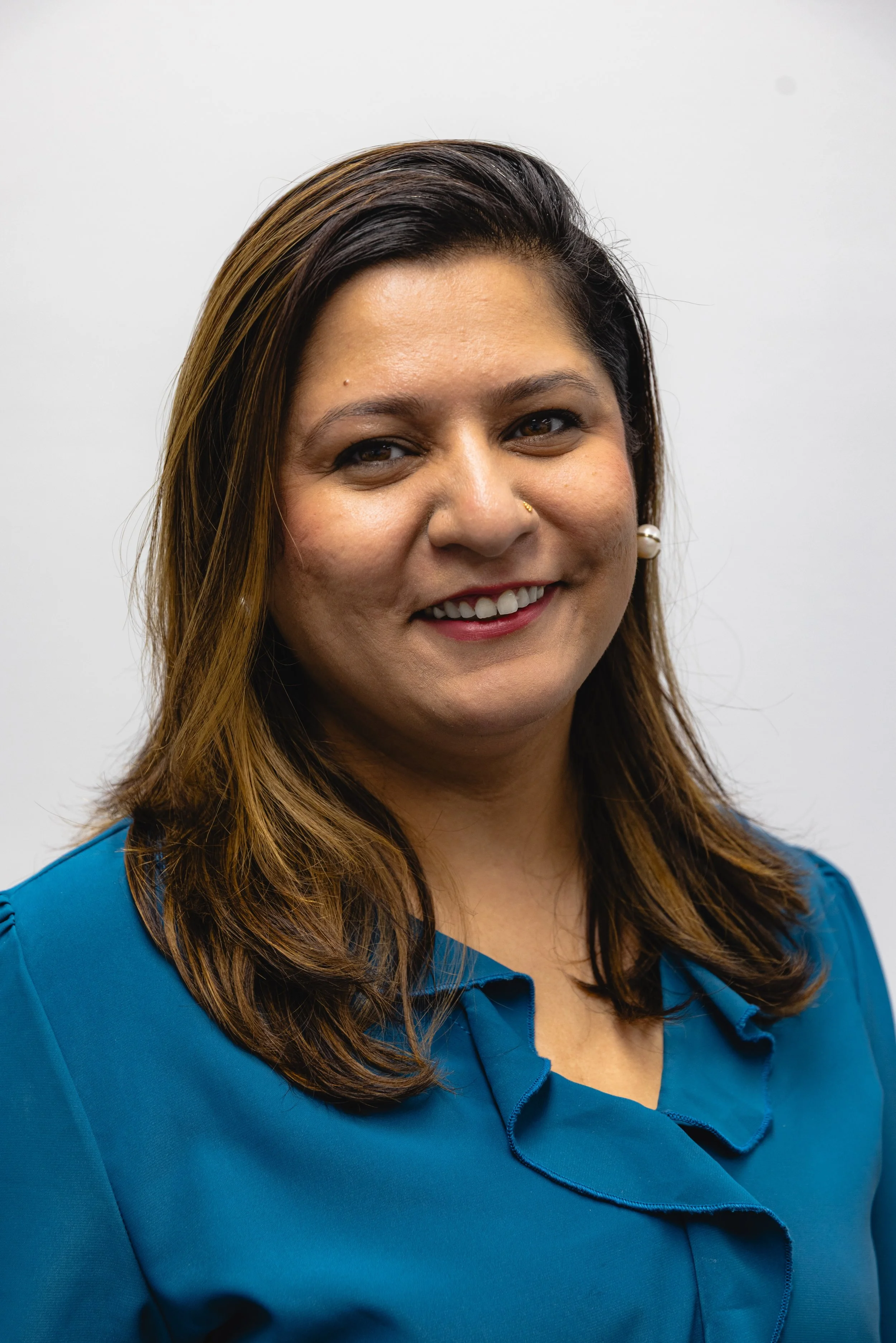Close-up of a smiling woman with long brown hair, wearing a blue blouse with ruffled collar, and a pearl earring, against a plain white background.