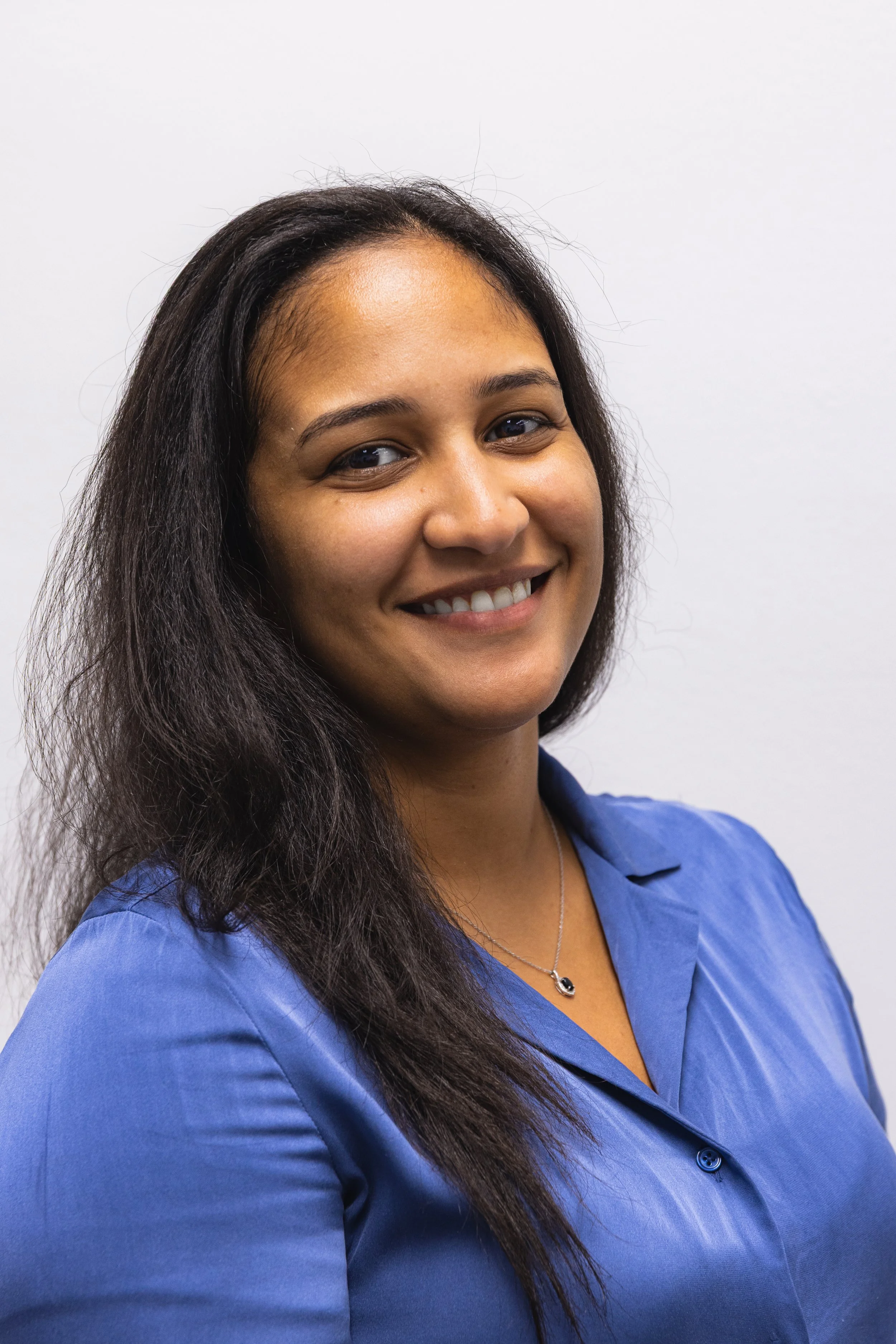 A smiling woman with long dark hair wearing a blue collared shirt and a silver necklace, standing against a plain white background.