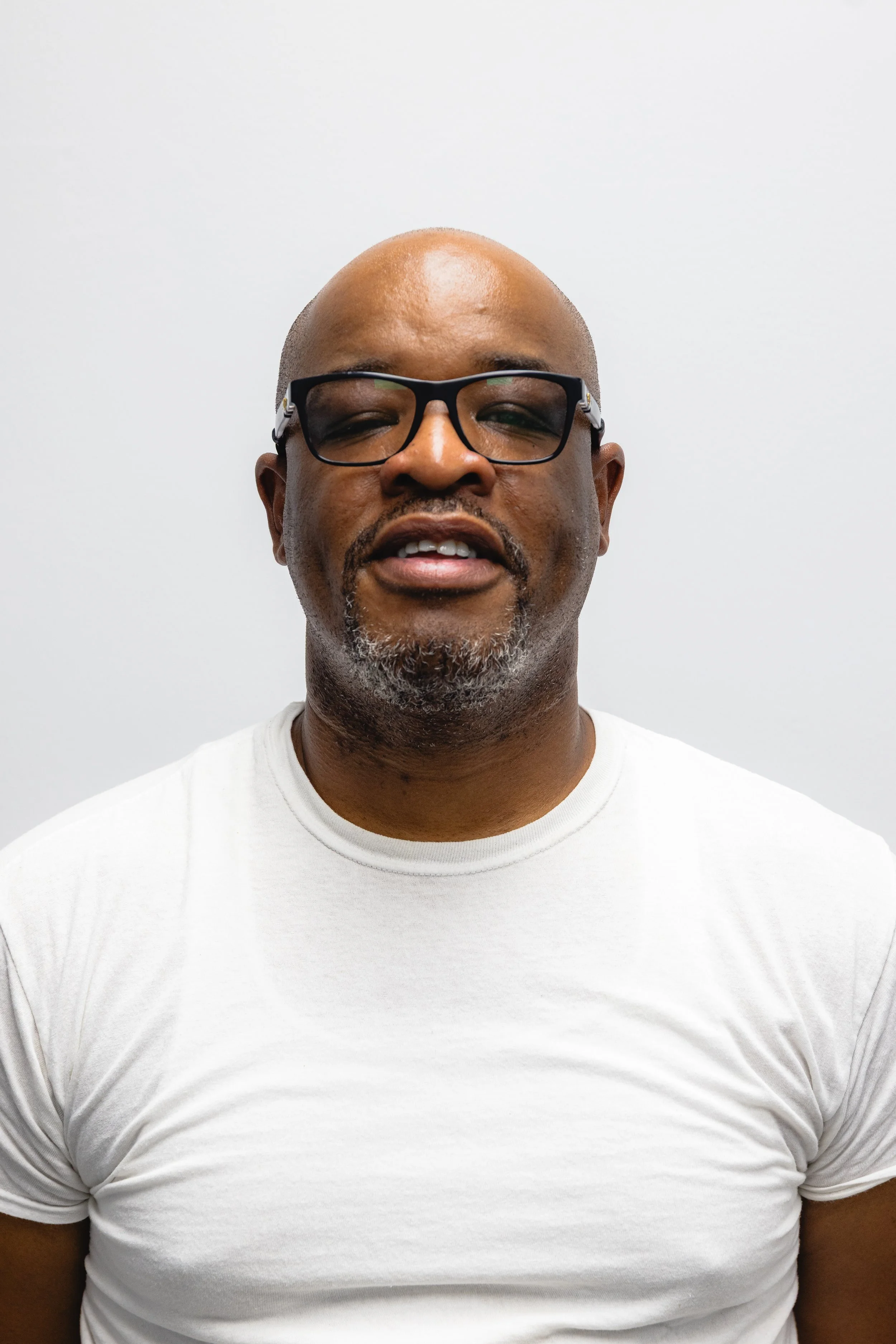 A man wearing glasses and a white t-shirt standing against a plain white wall.