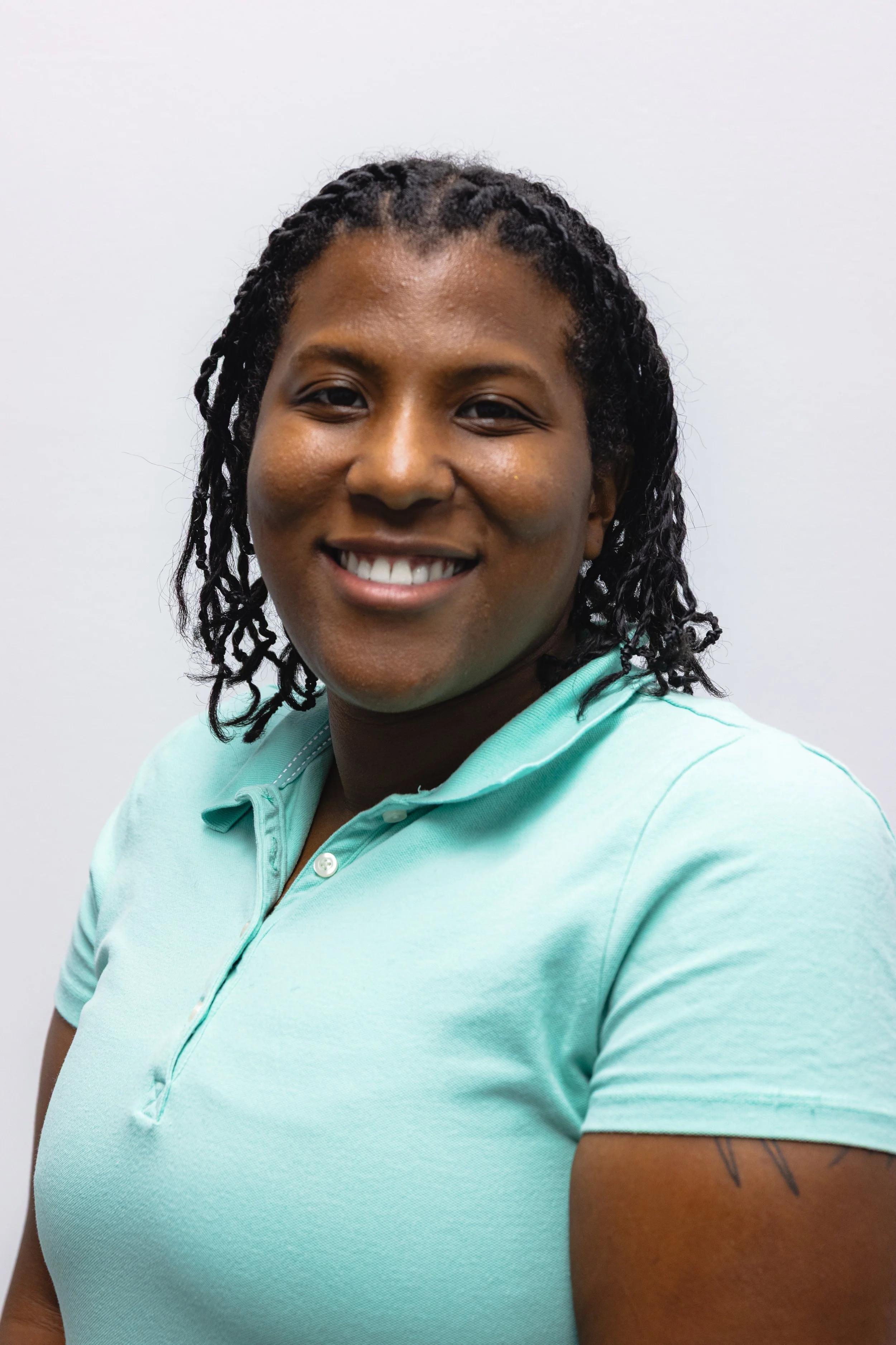 Close-up of a smiling woman with dark curly hair pulled back, wearing a light blue polo shirt, standing against a plain white background.