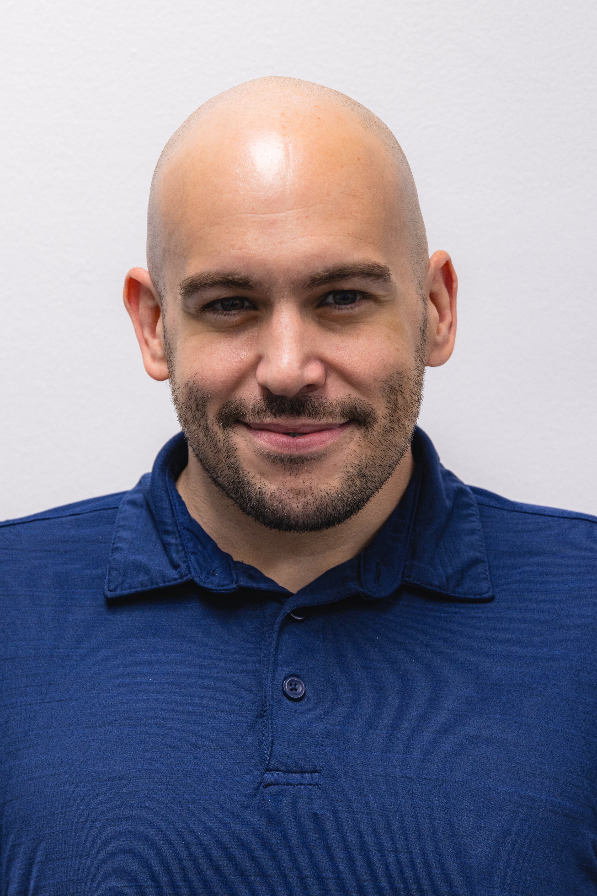 Close-up of a smiling man with a shaved head, beard, and blue eyes, wearing a blue button-up shirt, standing against a plain white wall.