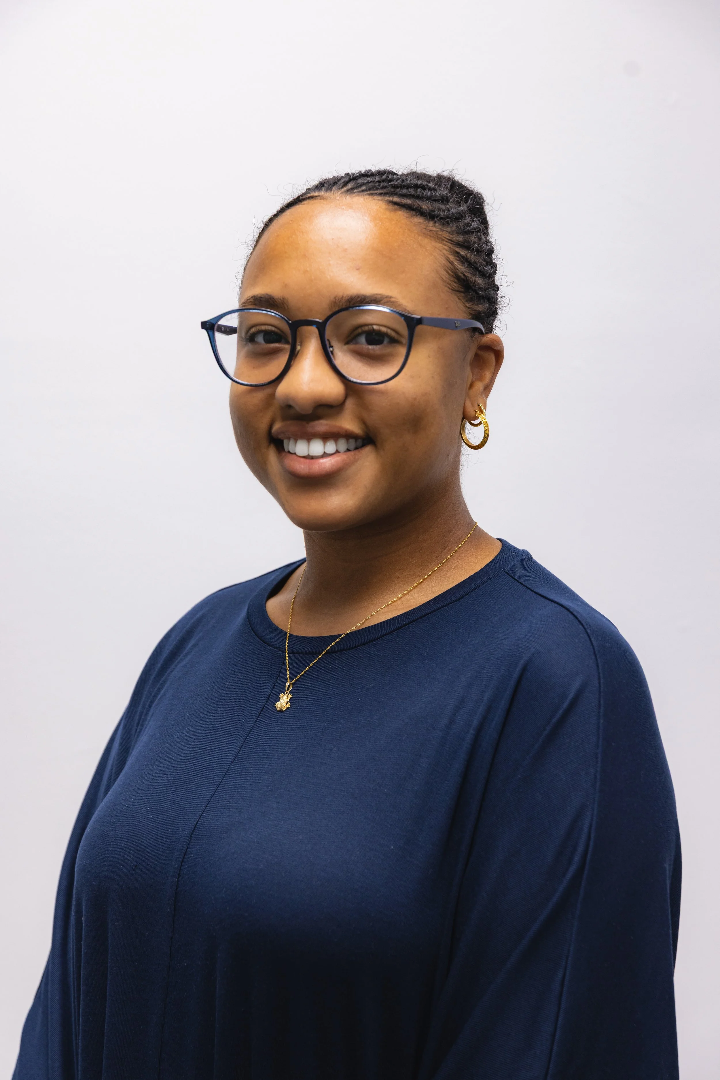 A young woman with glasses, gold earrings, and a necklace, smiling in front of a plain white wall.