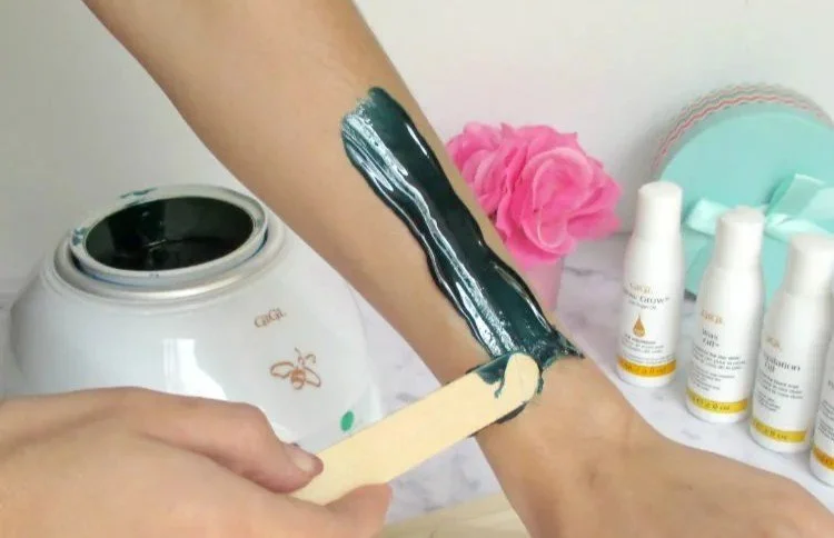 Person applying Gigi hard wax to their arm with a wooden spatula, with wax care products and a pink flower in the background.