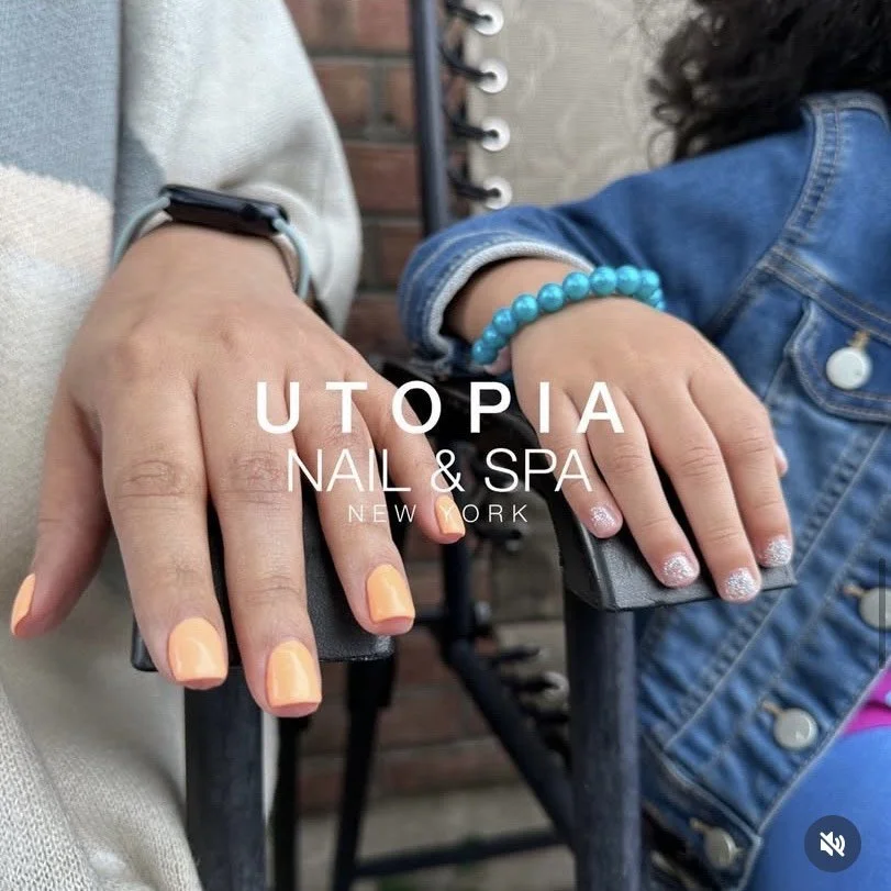 Close-up of two hands with freshly painted nails resting on black stands. One hand has peach-colored nails, and the other has silver-glitter nails. One person is wearing a smartwatch, and the other has a blue beaded bracelet. The background includes a brick wall and part of a chair, indicating a nail and spa salon setting. The text overlay reads 'UTOPIA NAIL & SPA NEW YORK.'