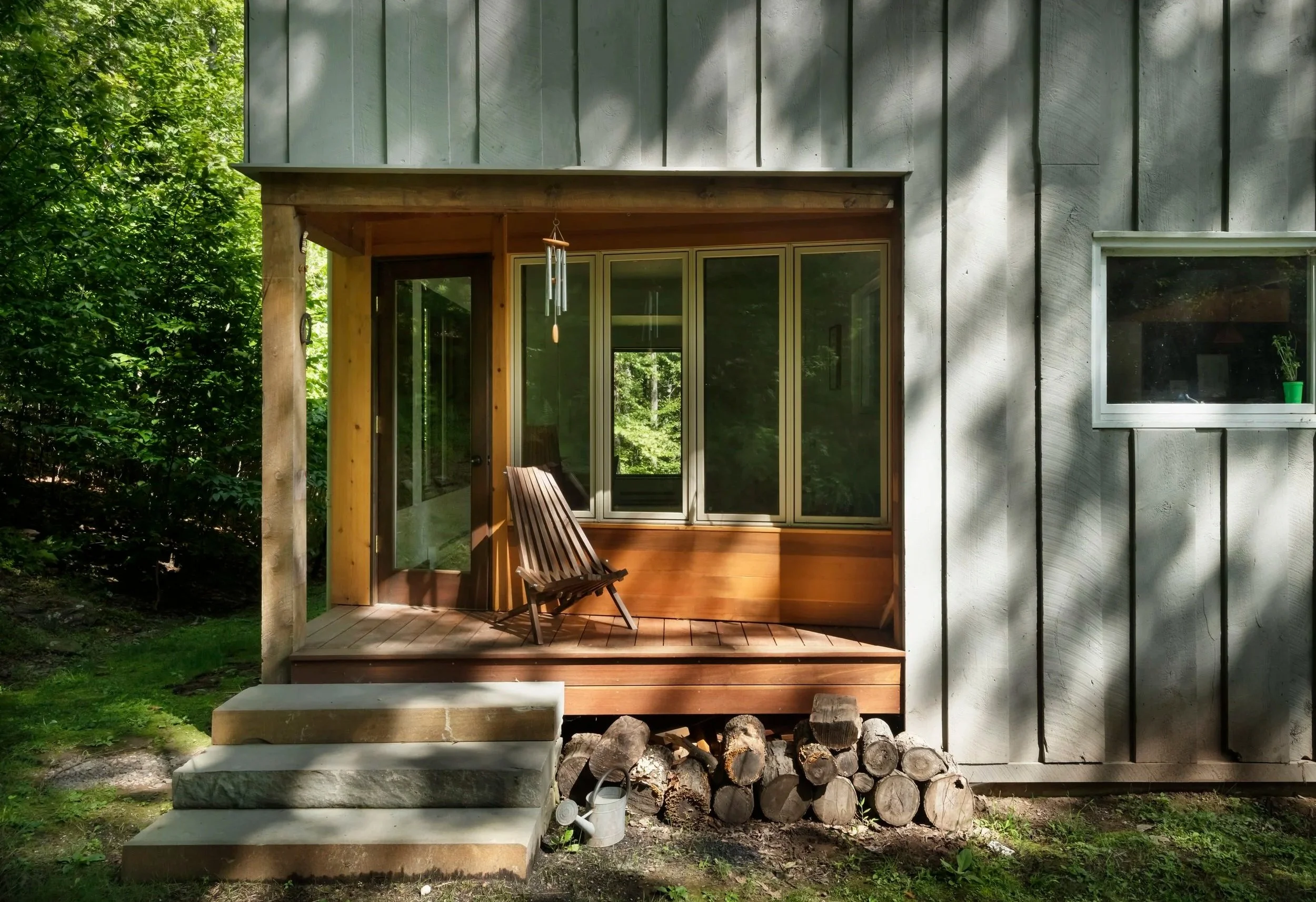 A small wooden porch with steps in front of a house with gray vertical siding, a window, and a sliding glass door, surrounded by green trees and grass.