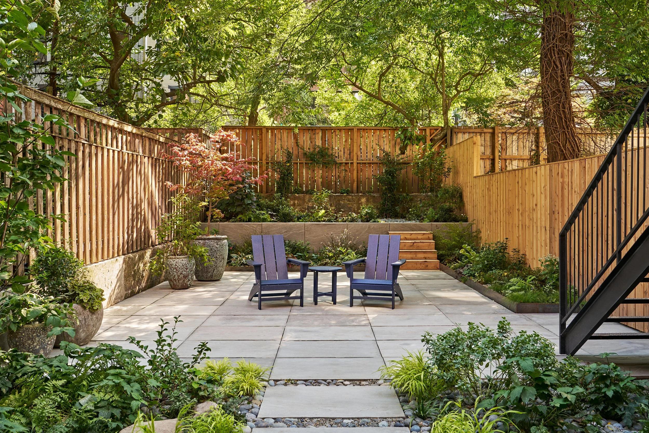 A backyard patio with two blue Adirondack chairs and a small table, surrounded by potted plants, greenery, and a wooden fence, with stairs leading to an elevated garden area and trees in the background.
