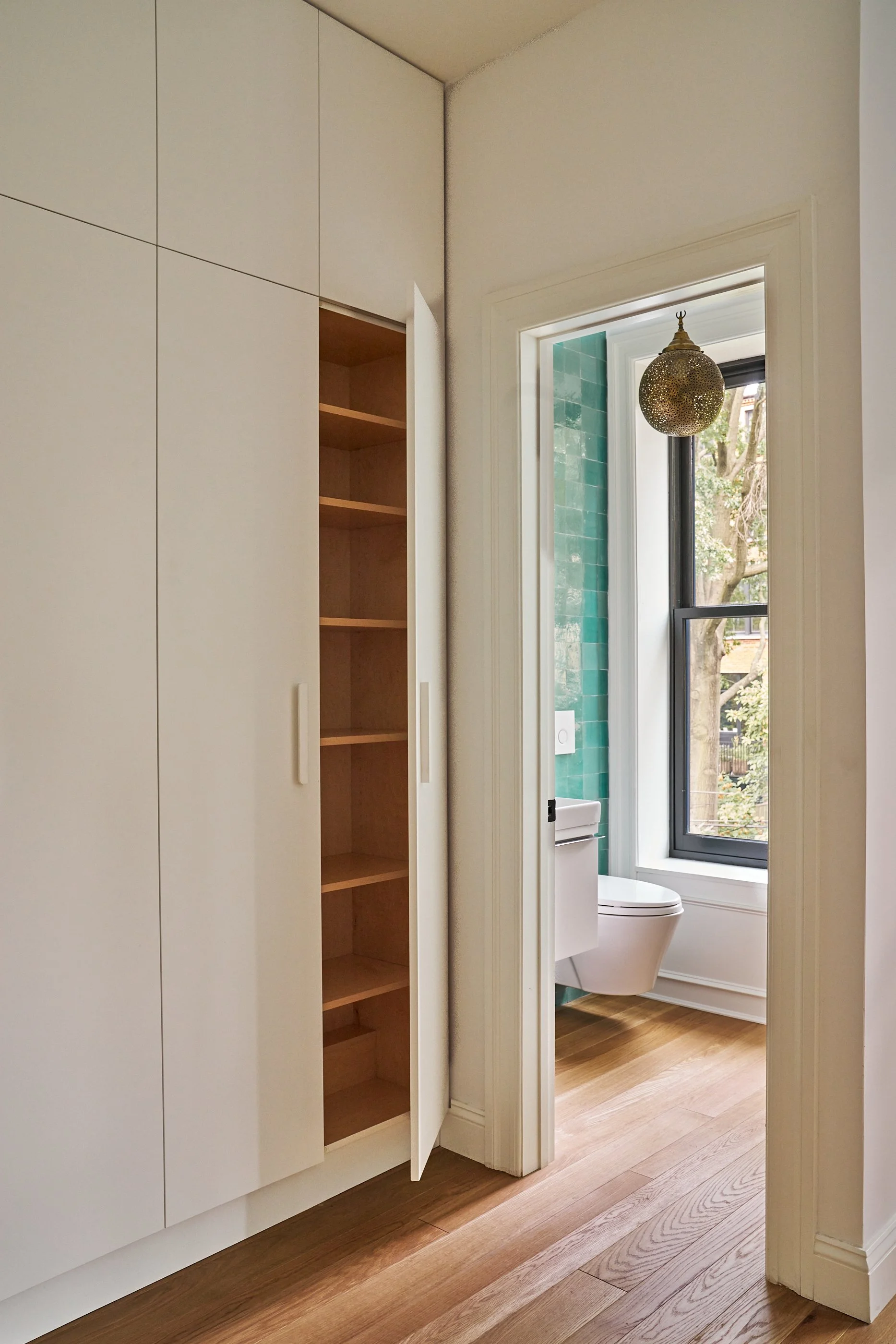 View of a hallway leading to a bathroom with a teal tiled accent wall. To the left, there is a built-in white cabinet with open light wood shelves inside. The bathroom has a large window, and a modern, wall-mounted toilet is visible beneath the window. A decorative hanging light fixture is also seen.