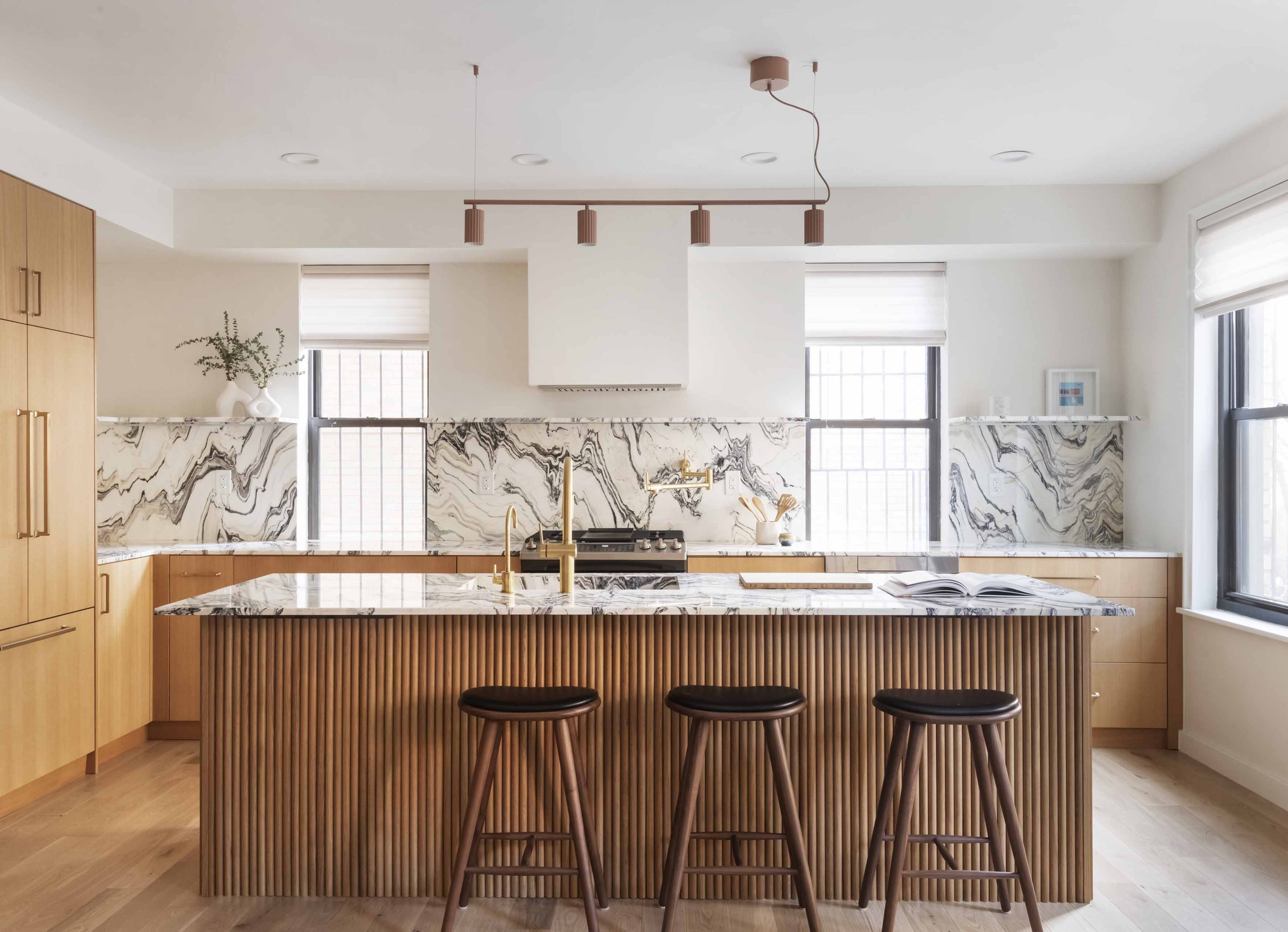 Modern kitchen with wooden cabinets, a marble island with a ribbed wooden front, black stools, gold fixtures, marble backsplash, and large windows.