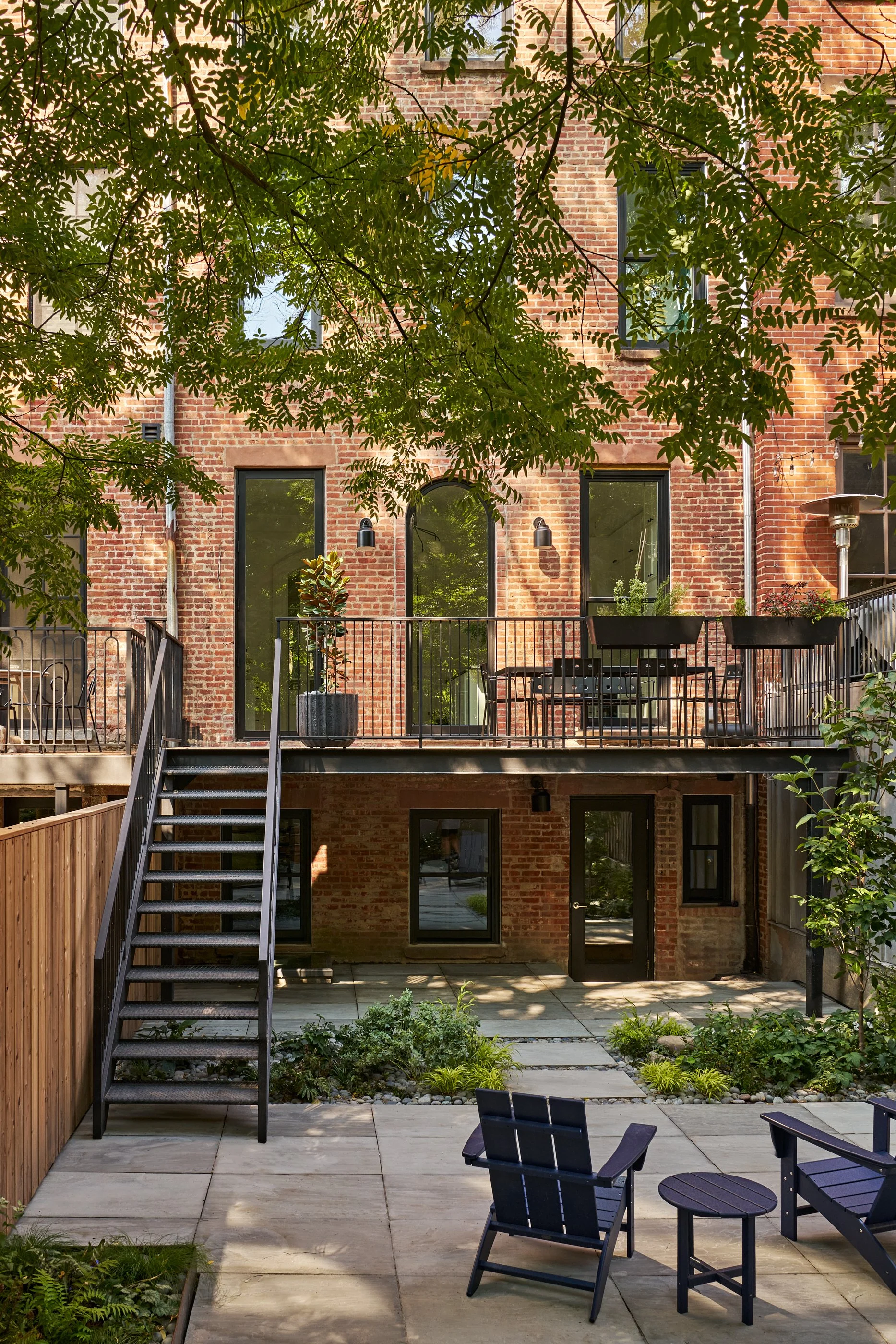 View of a brick apartment building with large windows, a small balcony with plants, and outdoor furniture on a lower patio, shaded by overhanging tree branches.