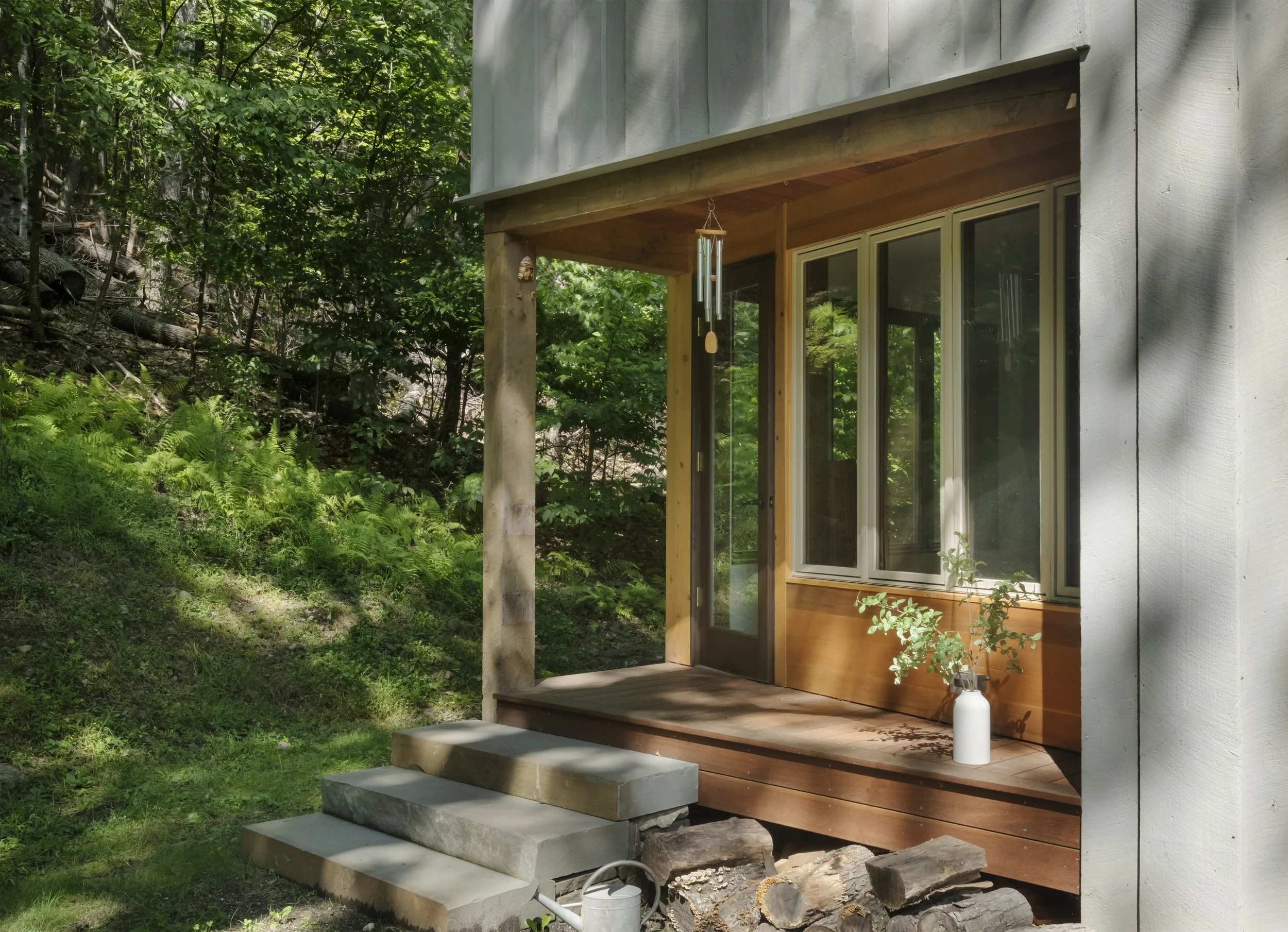 Front porch of a house with wooden steps, sliding glass door, and a white vase with green plant, surrounded by greenery and trees.