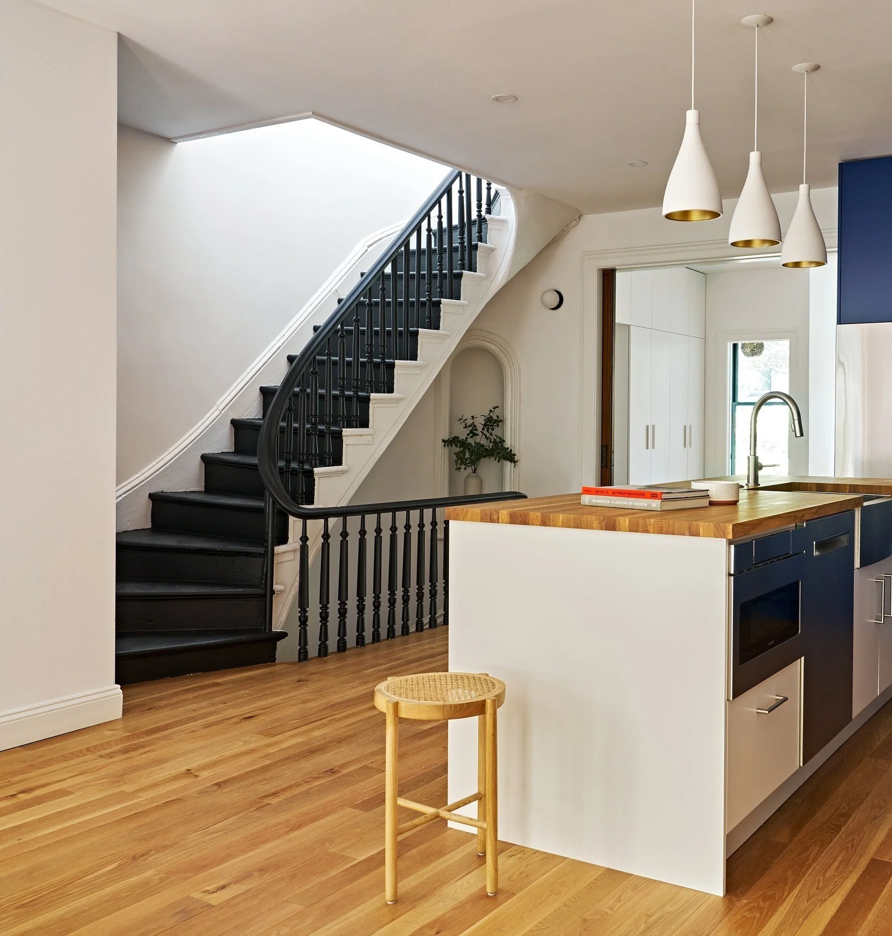 Modern kitchen with wooden floors, white walls, hanging pendant lights, black staircase with white risers, and a kitchen island with a wooden countertop, navy blue cabinets, a stainless steel oven, a faucet, and outdoor view through the window.