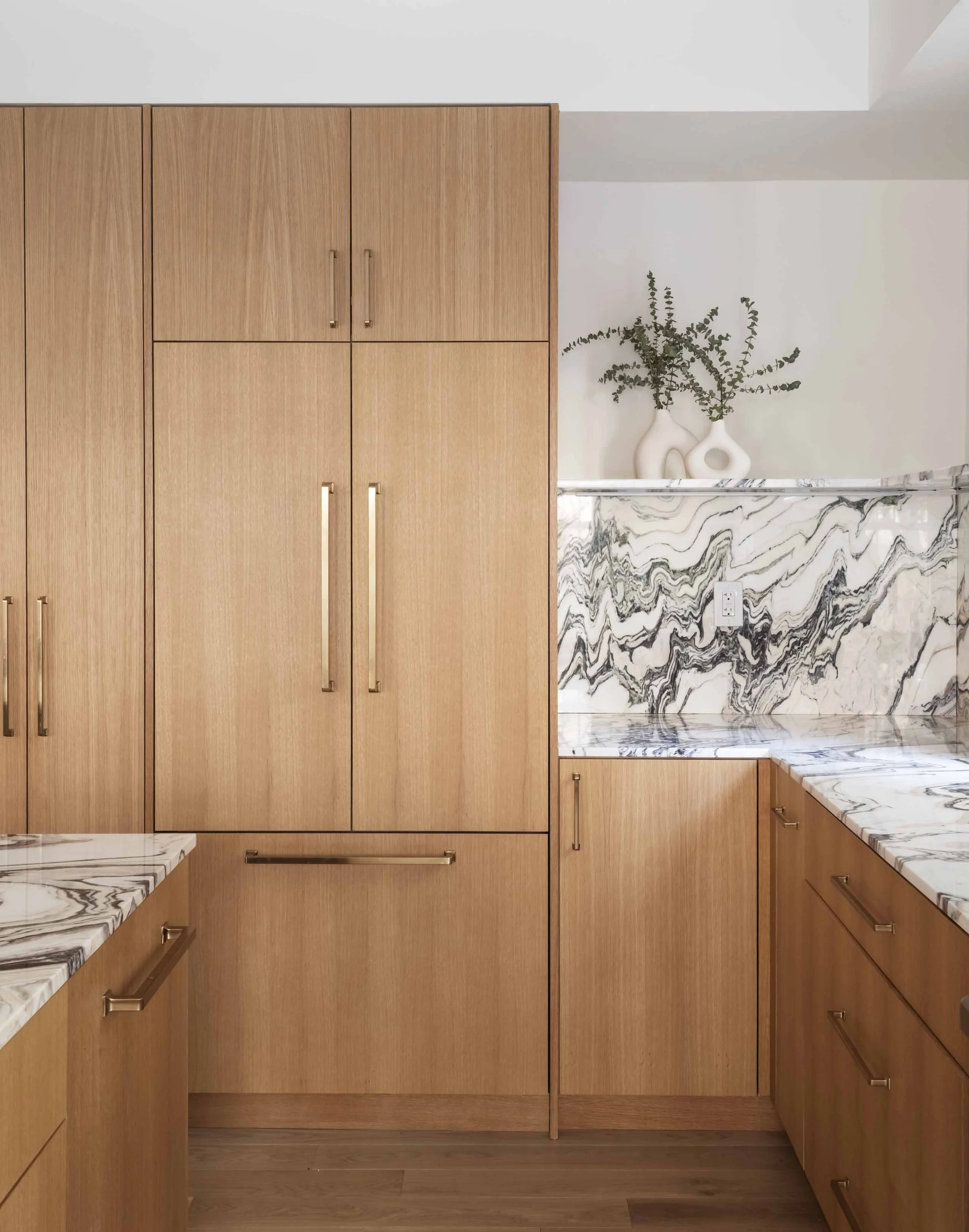 A kitchen with wooden cabinets, marble countertops, and a marble backsplash; two white vases with green plant on the counter.