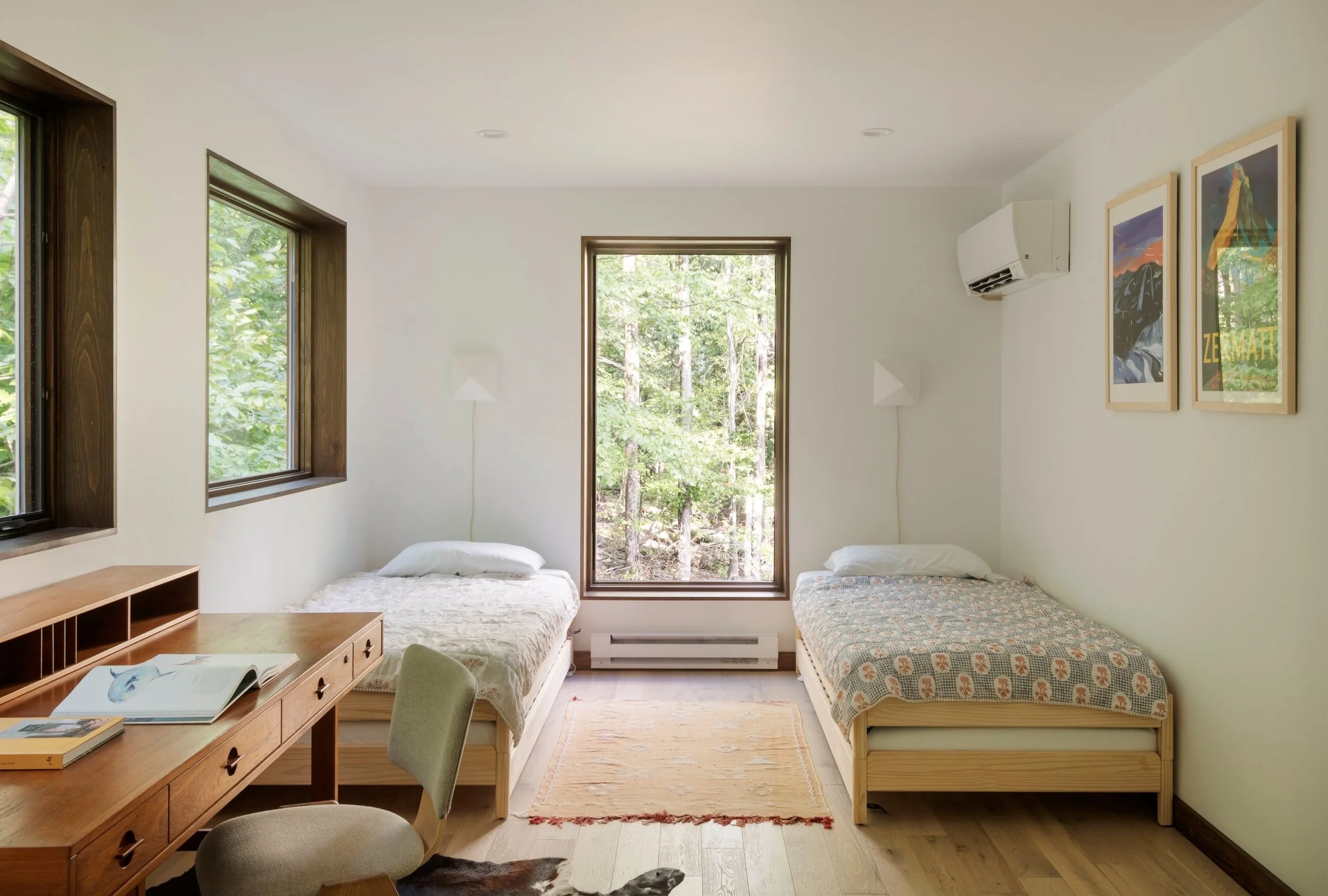 A bedroom with two beds near large windows showing greenery outside, a wooden desk with an open book, framed art on the wall, and an air conditioning unit.