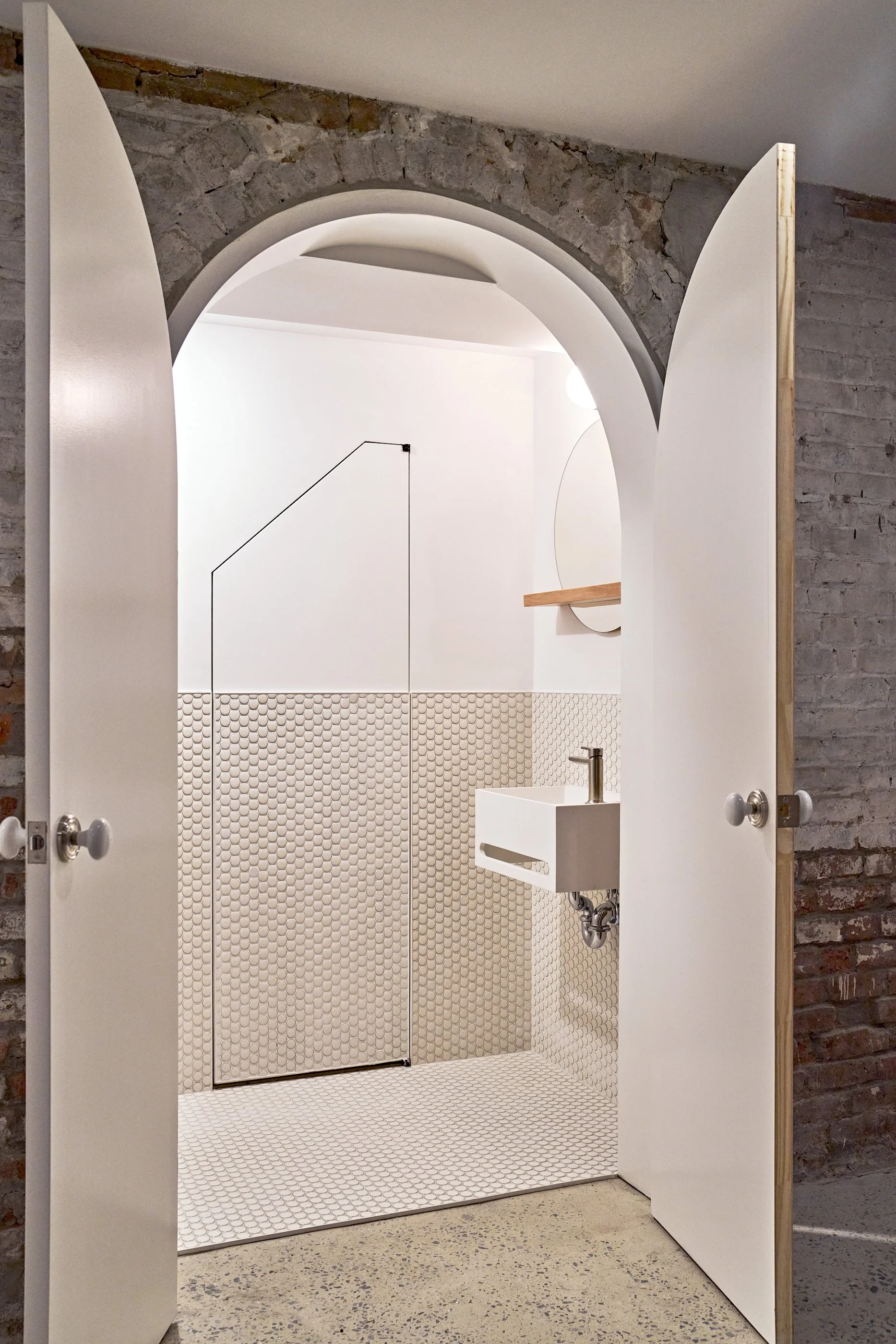 A modern, minimalist bathroom with white walls and a small white sink, viewed through arched double doors. The bathroom features textured beige tile on the lower half of the walls and floor, and an exposed brick archway at the doorway.