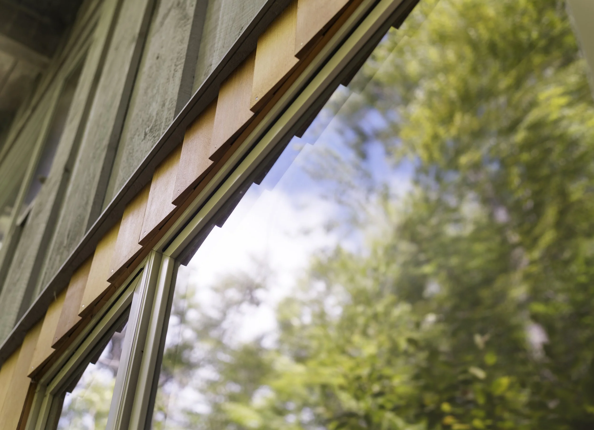 Close-up of a window with wooden and metal components, reflecting green trees and blue sky outside.