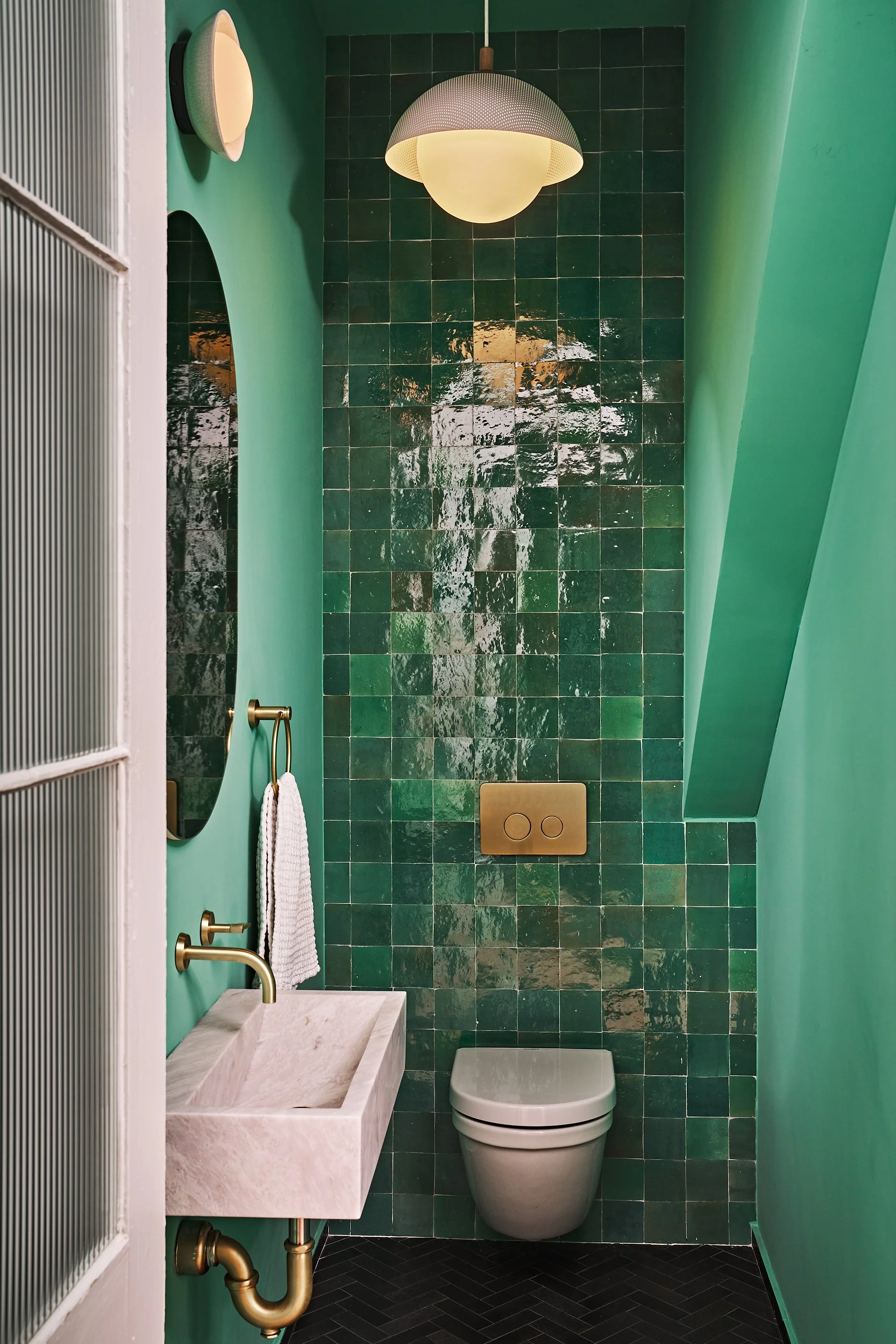 Small modern bathroom with green tiled wall behind a wall-mounted white toilet, a beige marble sink with gold fixtures, a gold towel ring with a white towel, a round mirror, and a ceiling light fixture. The walls are painted green and the floor has black herringbone tiles.