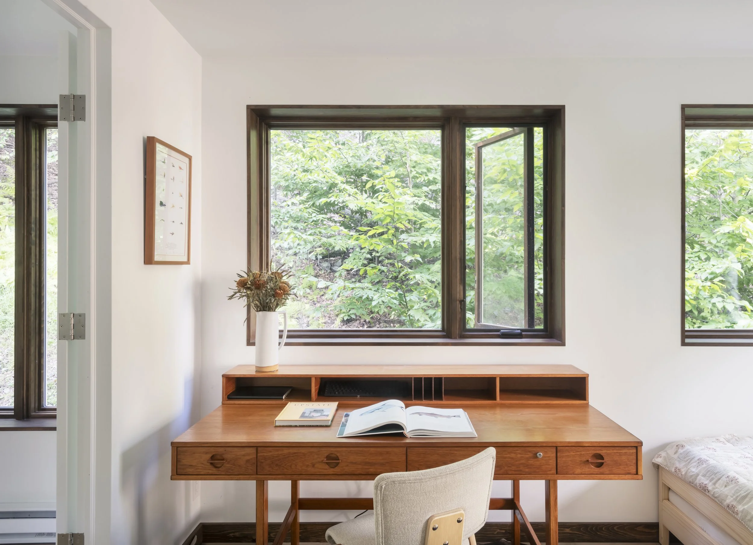 A cozy study space with a wooden desk, open book, and a chair, illuminated by large windows showing green foliage outside.