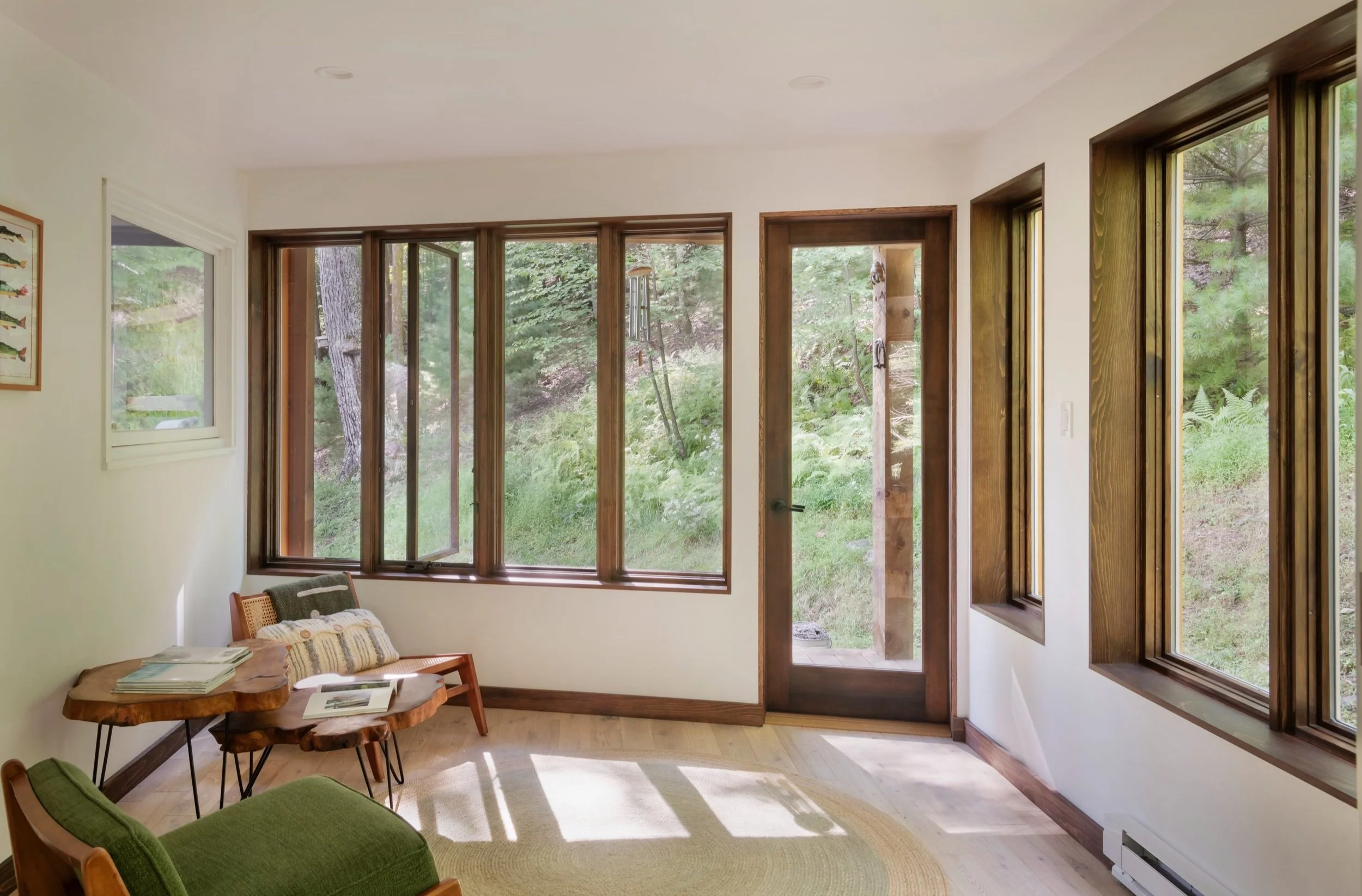 Sunlit living room with large windows and a door opening to a wooded outdoor area. Elm and wooden furniture, including a green armchair, a wood table, and a bench, are arranged in the room.