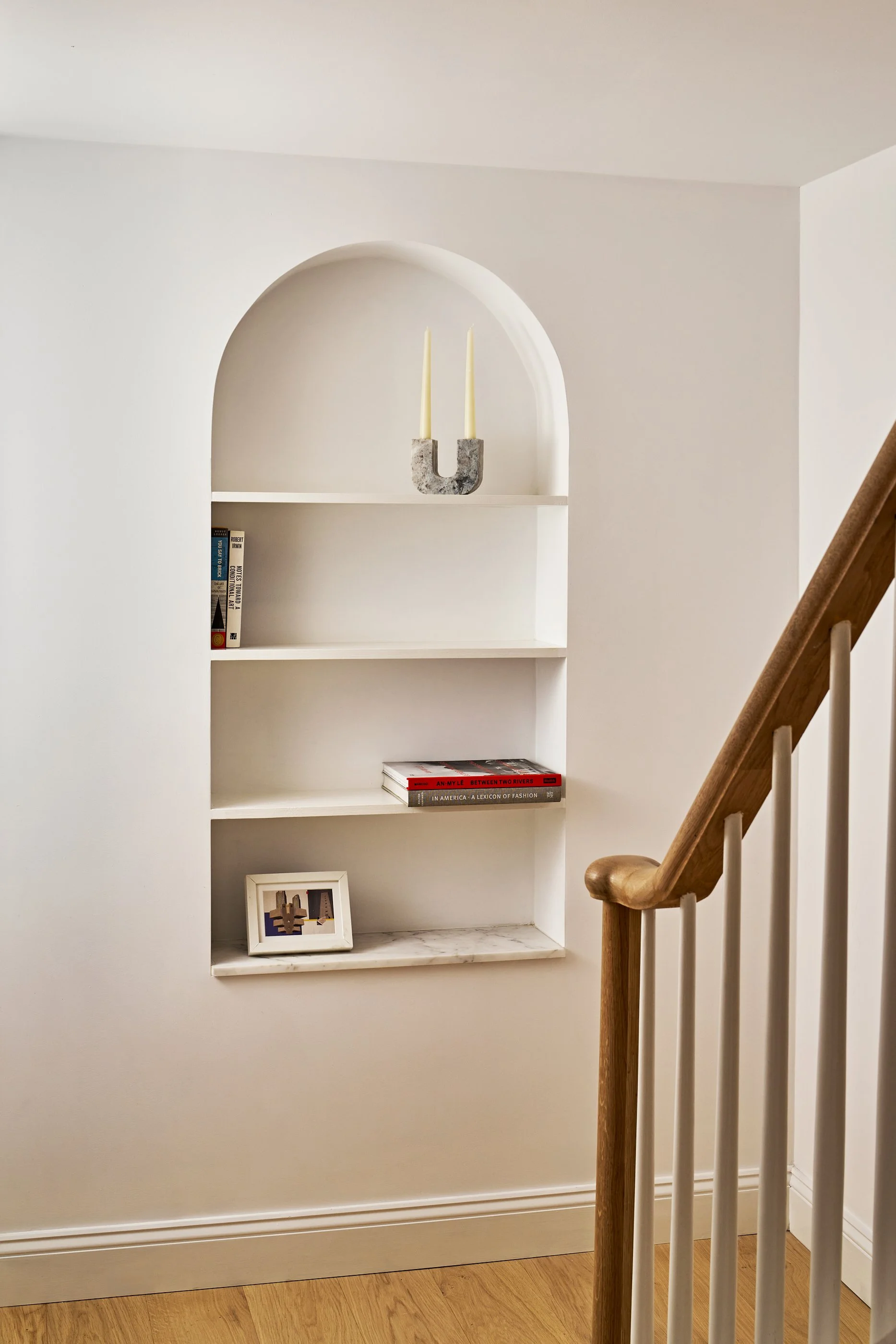 Built-in white wall nook with three shelves, decorative artist's candle holder with two candles on the top shelf, books and framed pictures on the middle and lower shelves, and a wooden staircase railing to the right.