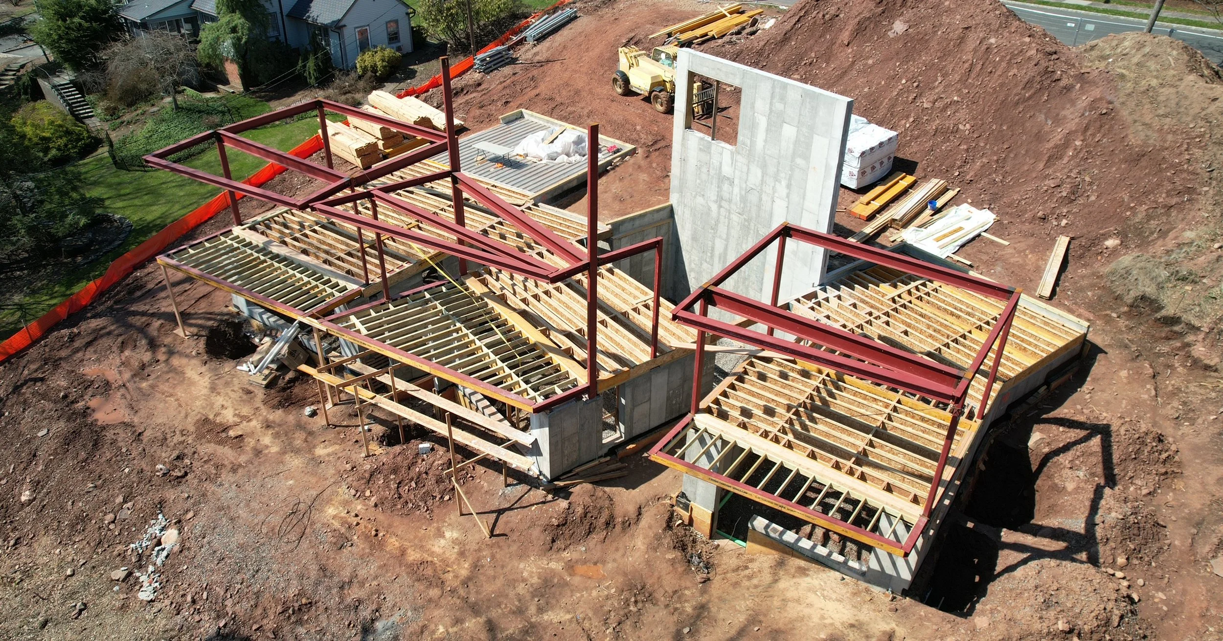 Construction site with framing for a building, steel beams, and a concrete wall. Construction materials and equipment are visible, with a green lawn and trees in the background.