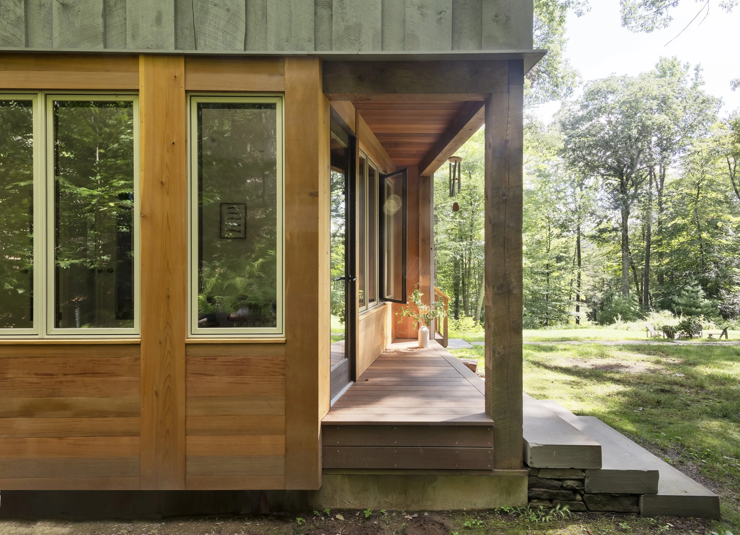 Front porch of a wooden house with open windows, a hanging wind chime, and a potted plant, set amidst a lush green forest.