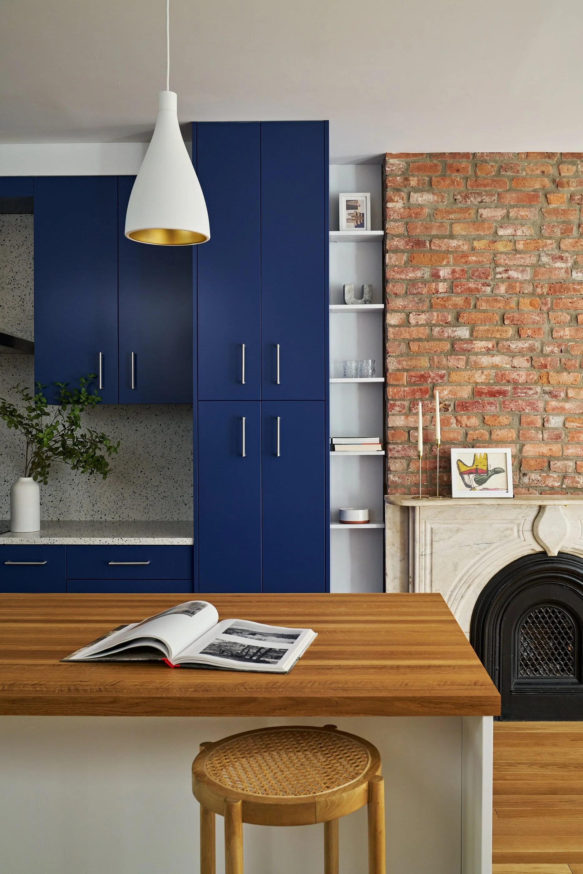 Interior of a modern kitchen with blue cabinets, a wooden table, a white pendant light, a brick fireplace, and decorative items.