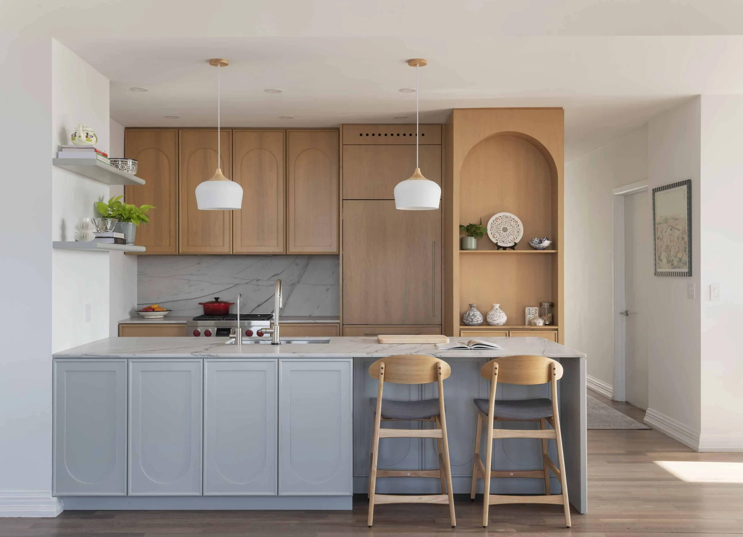 Modern kitchen with a blue island, wooden cabinets, white shelves with books and decor, pendant lights, a marble backsplash, and a breakfast bar with wooden chairs.